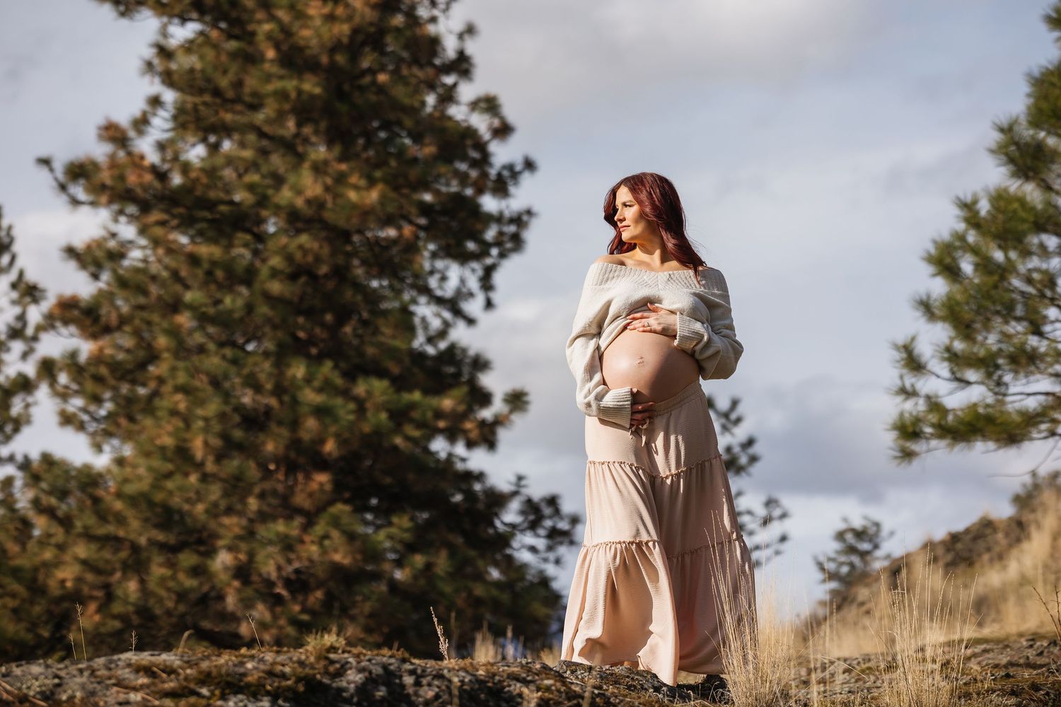 A woman in a flowing cream dress stands gracefully in a golden field with pine trees in the background at sunset.