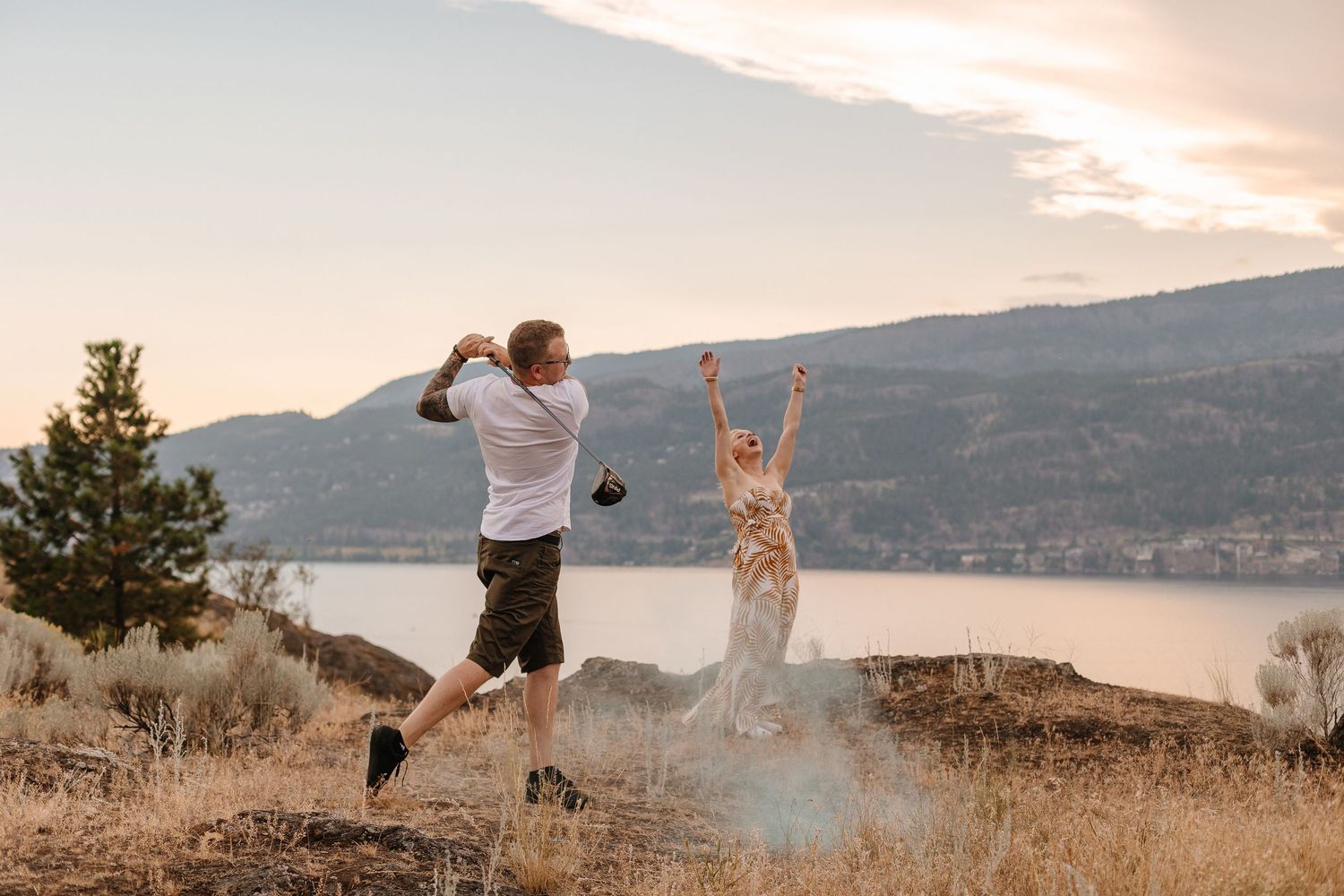 Two people jumping joyfully on a hilltop overlooking a lake at sunset with mountains in the background.