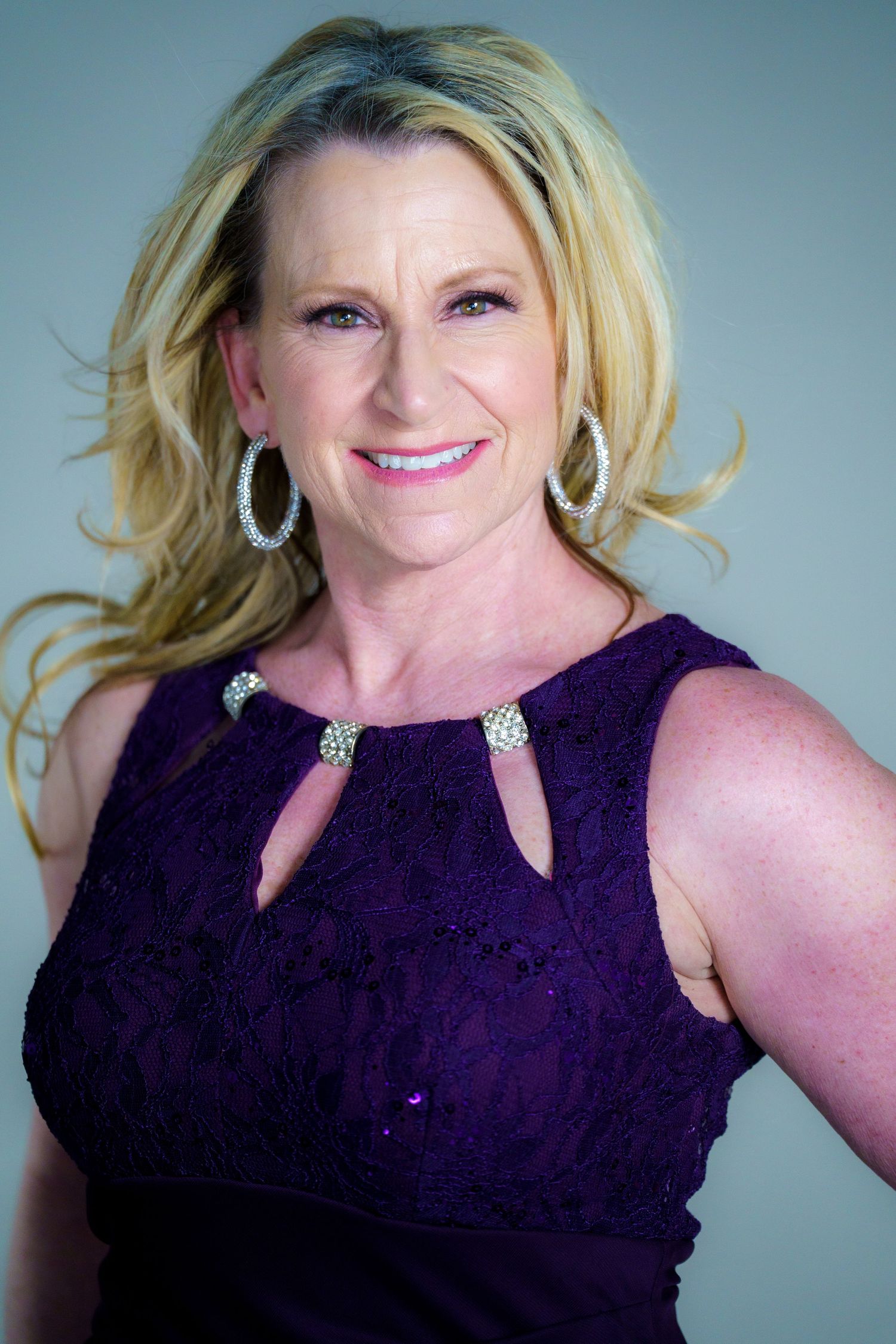 Professional headshot featuring a person in a purple sleeveless dress with cutouts and sparkly hoop earrings against blue backdrop.