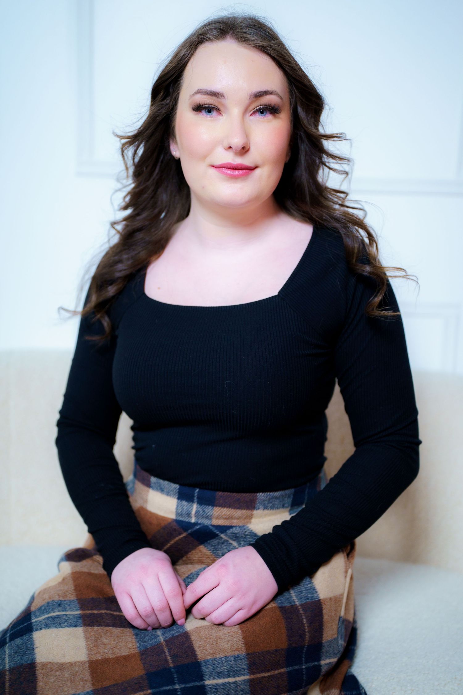 Professional portrait in a black long sleeve top and plaid skirt sitting against light colored backdrop.