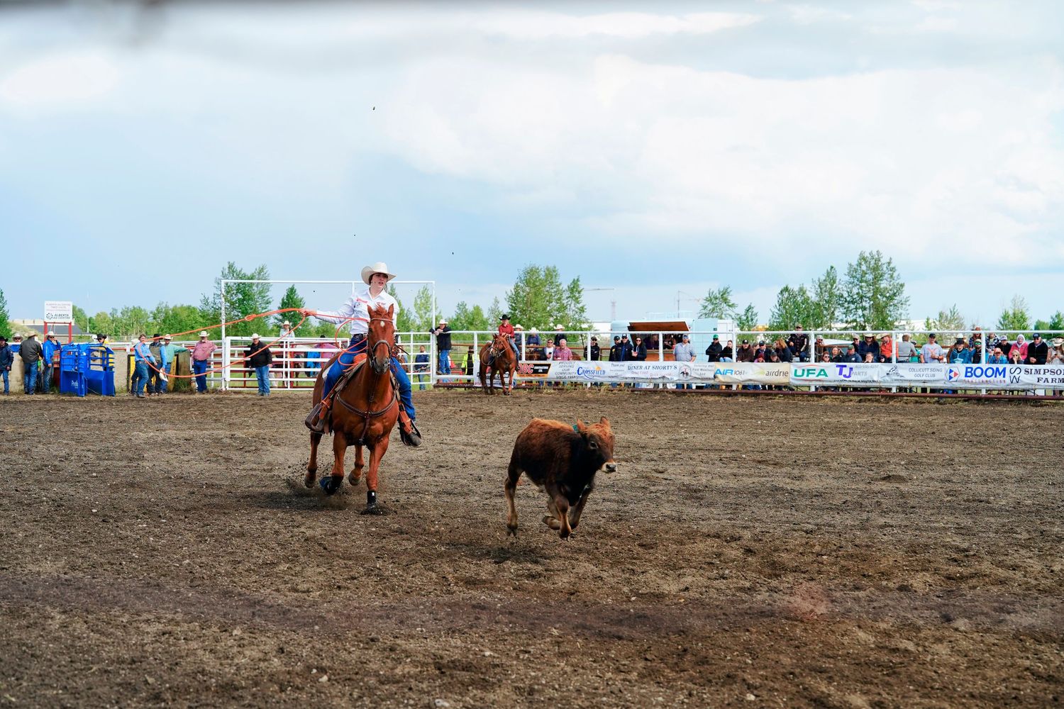 Cowboys compete in a fast-paced roping event at a rodeo arena with spectators watching from the sidelines.