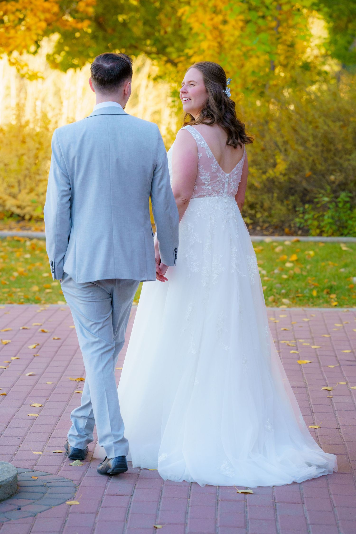Newlywed couple walking together on brick path surrounded by fall foliage in elegant wedding attire.