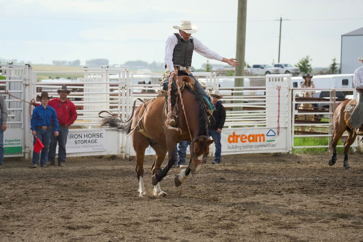A rodeo competitor on horseback performs in a dirt arena with spectators and white fencing visible in the background.