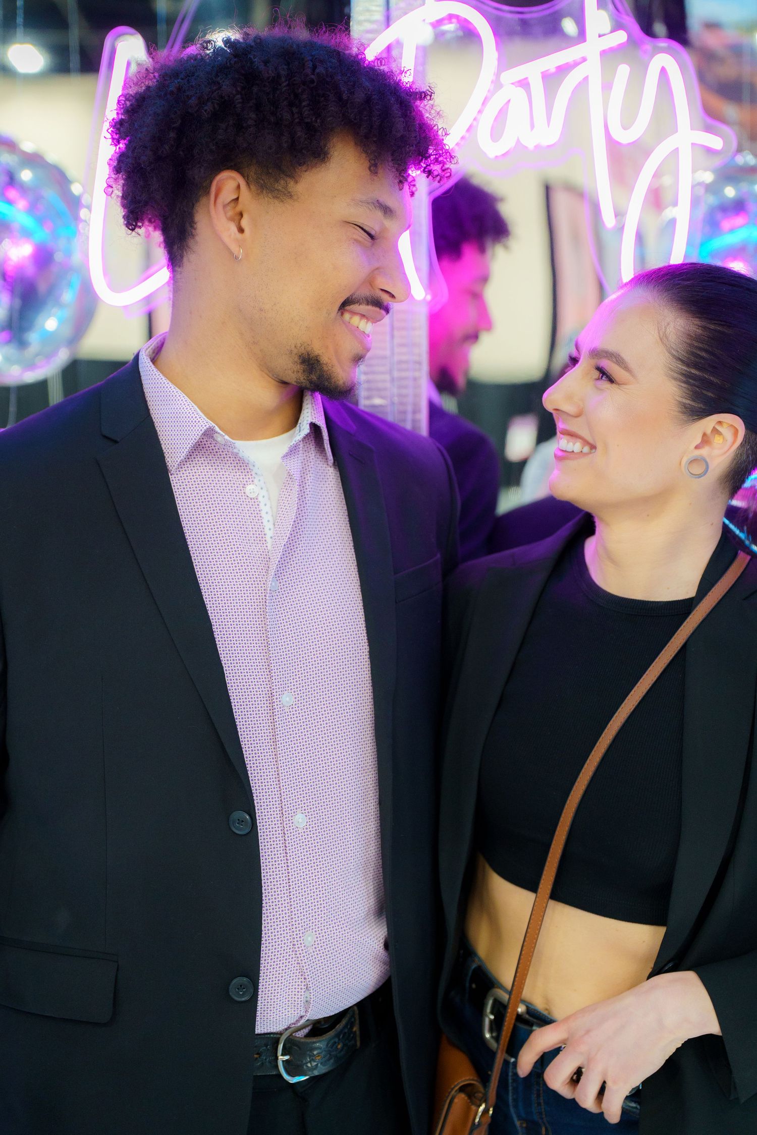 Couple smiling at each other in front of neon lights, with person wearing black suit and pink shirt and another in black top.