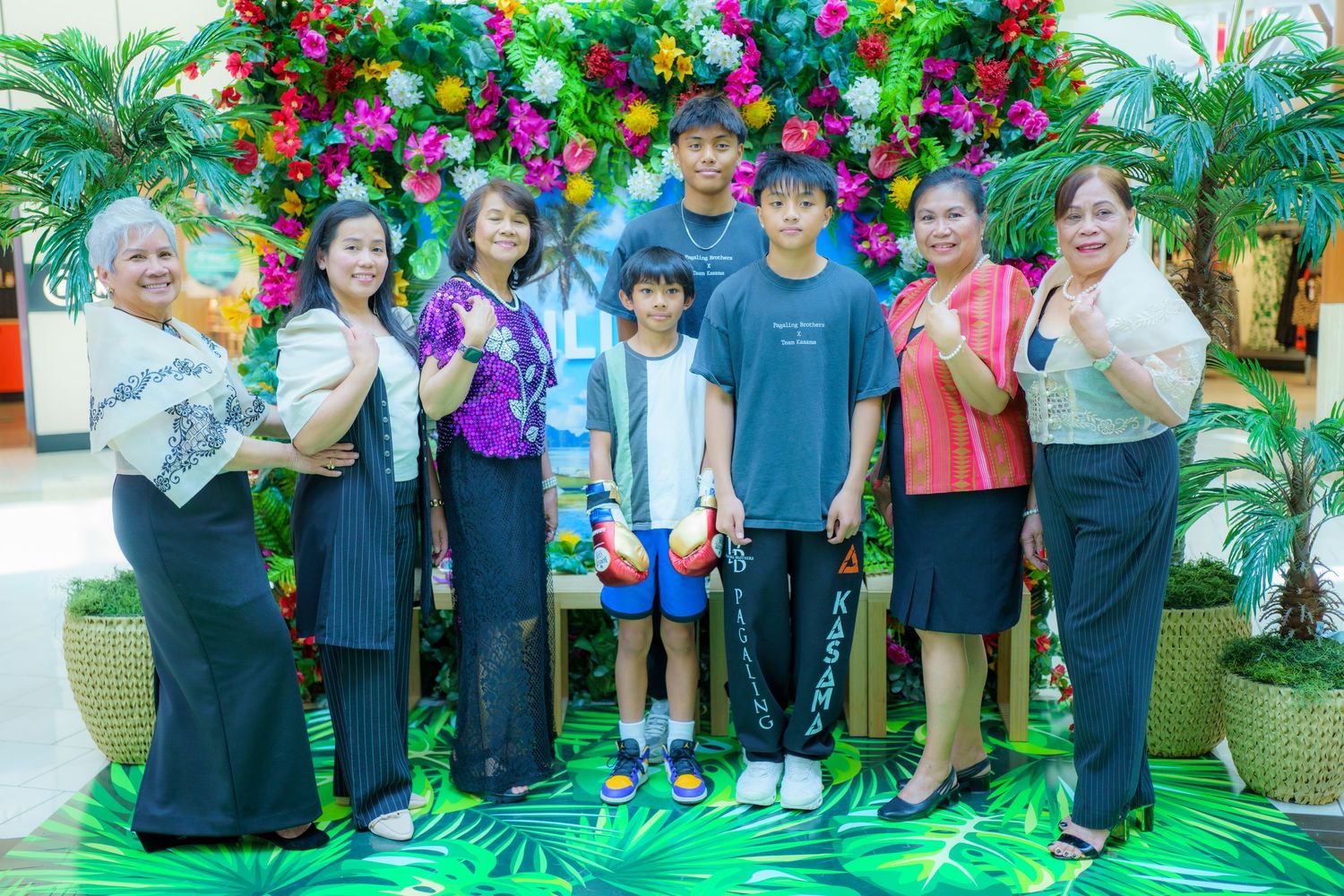 A group poses together in front of a tropical backdrop with colorful flowers and palm leaves.