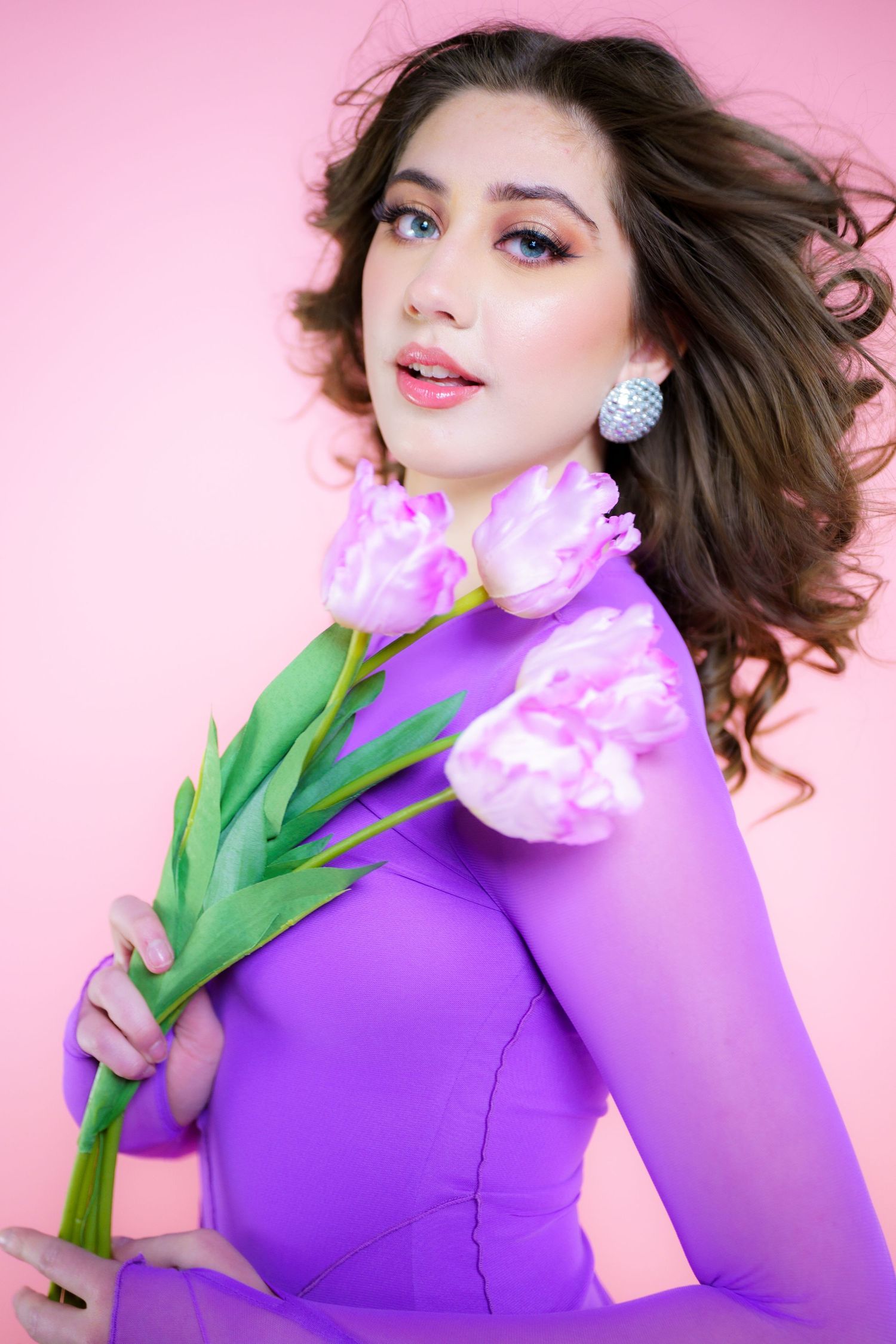 Portrait against pink backdrop showing flowing hair, bright blue eyes, purple dress and pink tulip flowers held elegantly.