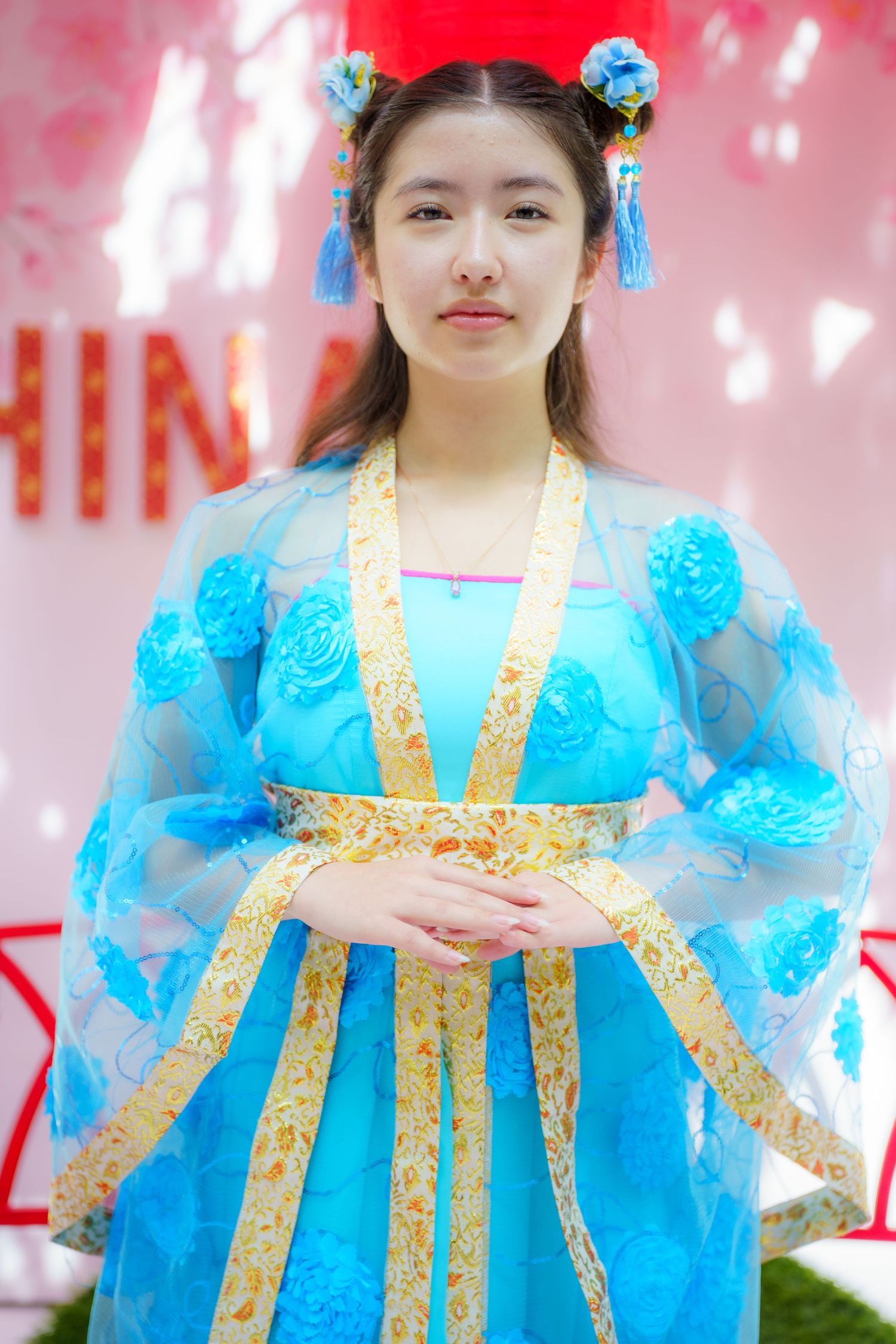 Traditional Chinese hanfu costume in bright turquoise blue with gold trim and floral hair accessories against pink background.