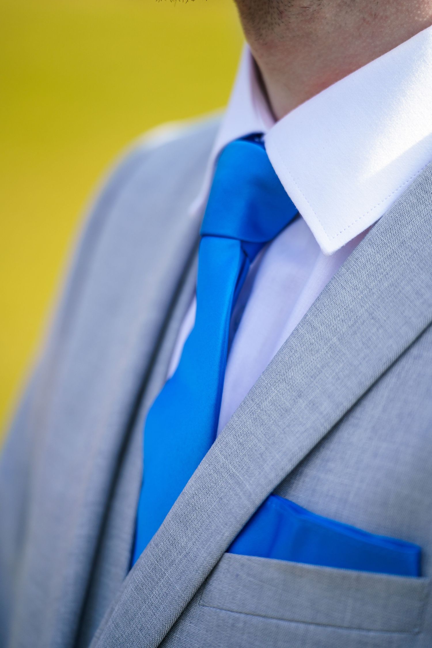 Close-up detail of a formal gray suit with bright blue tie and matching pocket square against yellow background.