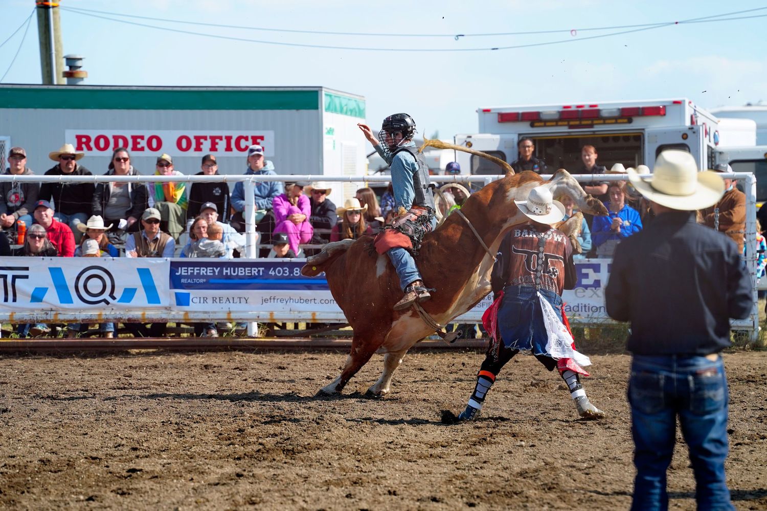 Rodeo bull riding competition with a cowboy competing in front of a crowd at an outdoor arena during daytime.