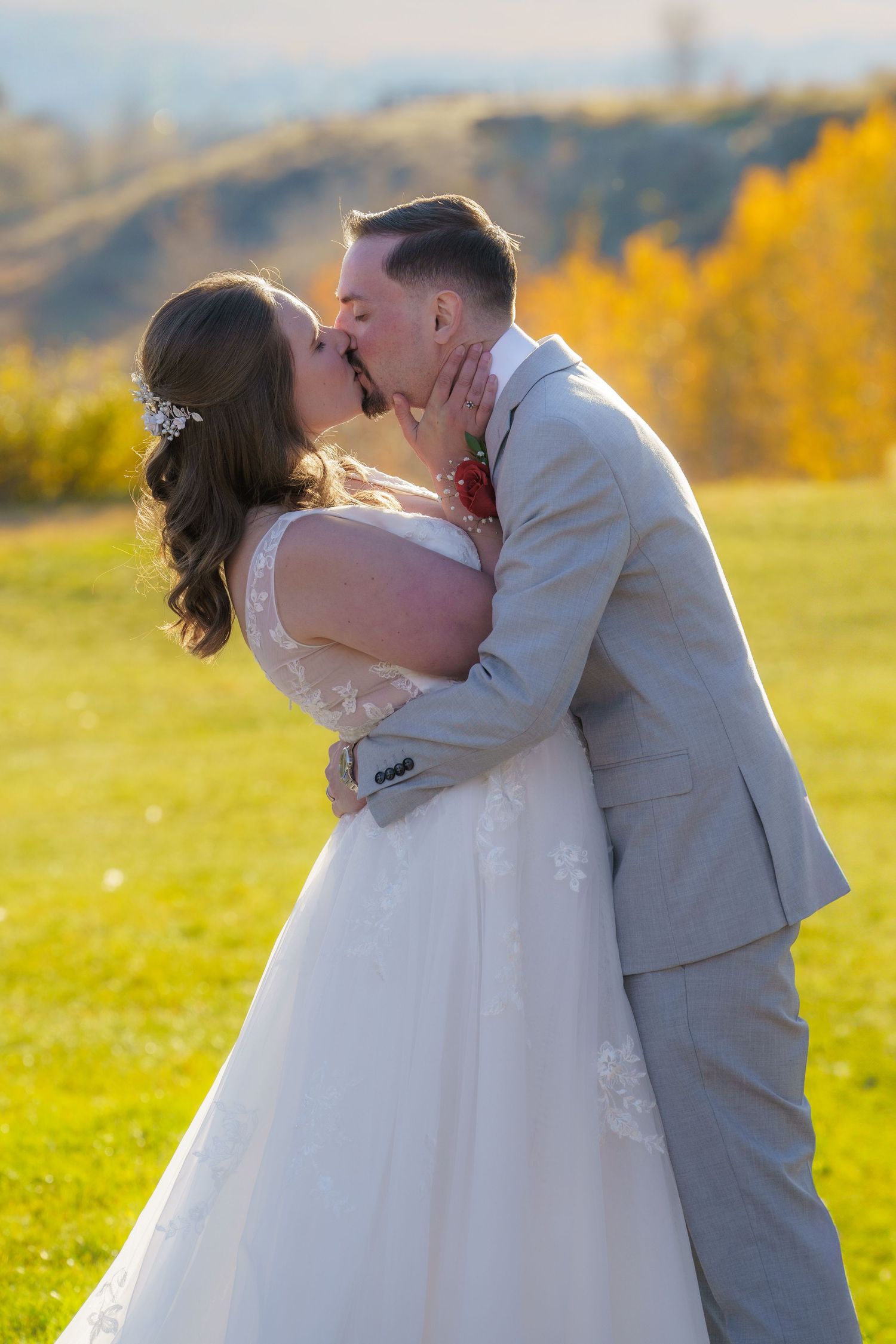 A couple shares their first kiss as newlyweds in a scenic mountain setting with golden autumn foliage in the background.