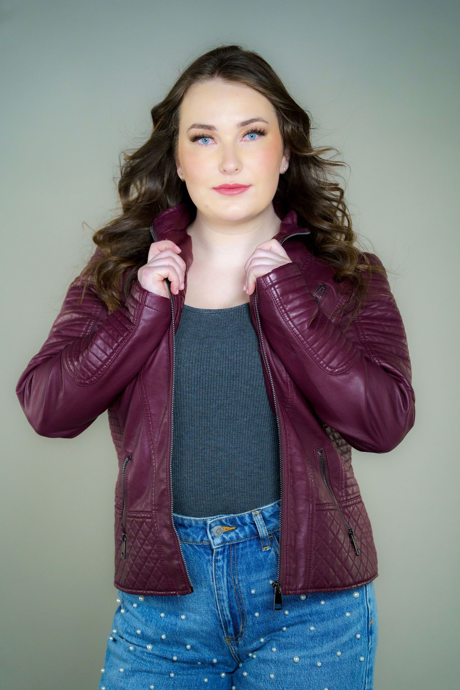 Casual fashion portrait with burgundy leather jacket, blue jeans, and flowing brown hair against gray background.