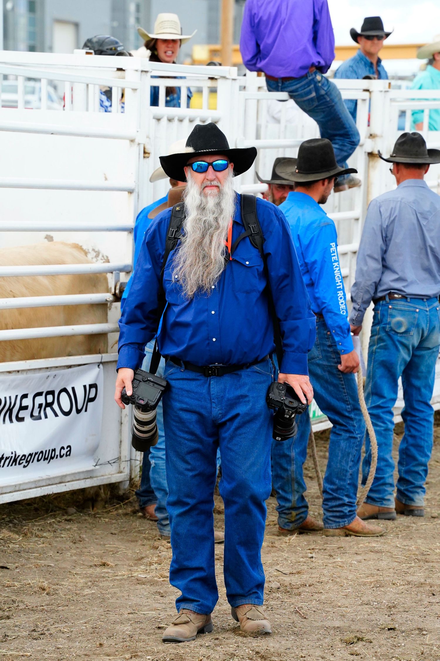 Professional photographer in blue western attire with long gray beard and sunglasses stands at rodeo grounds holding camera gear.