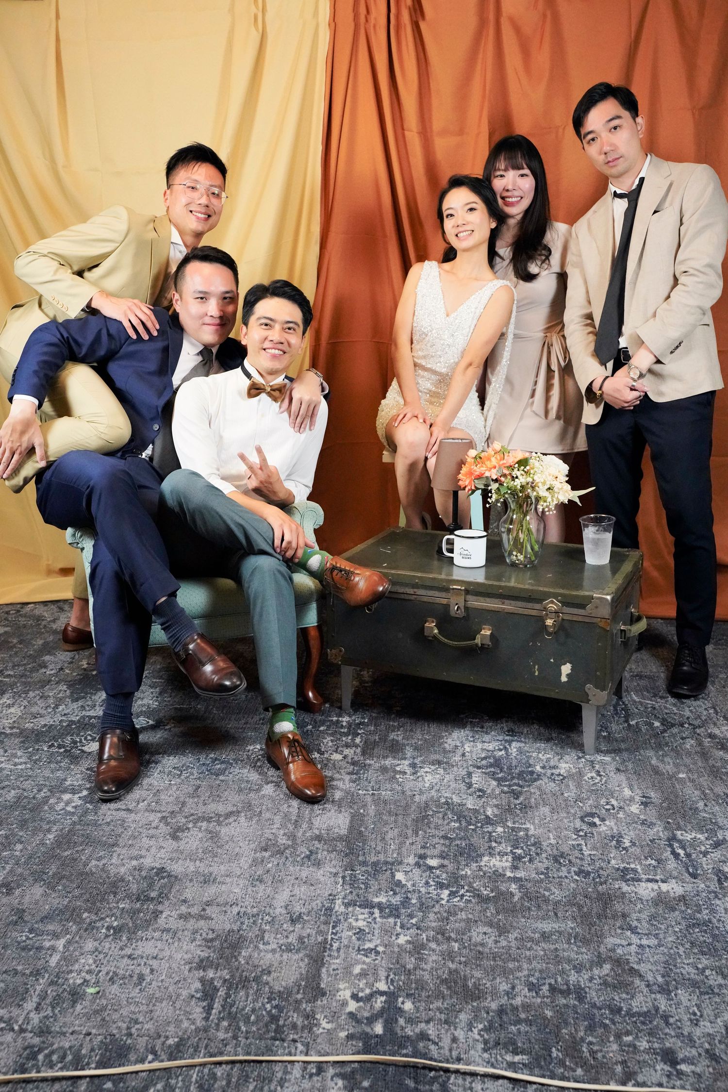 A group in formal attire poses together against a draped fabric backdrop with a vintage suitcase and floral arrangement.