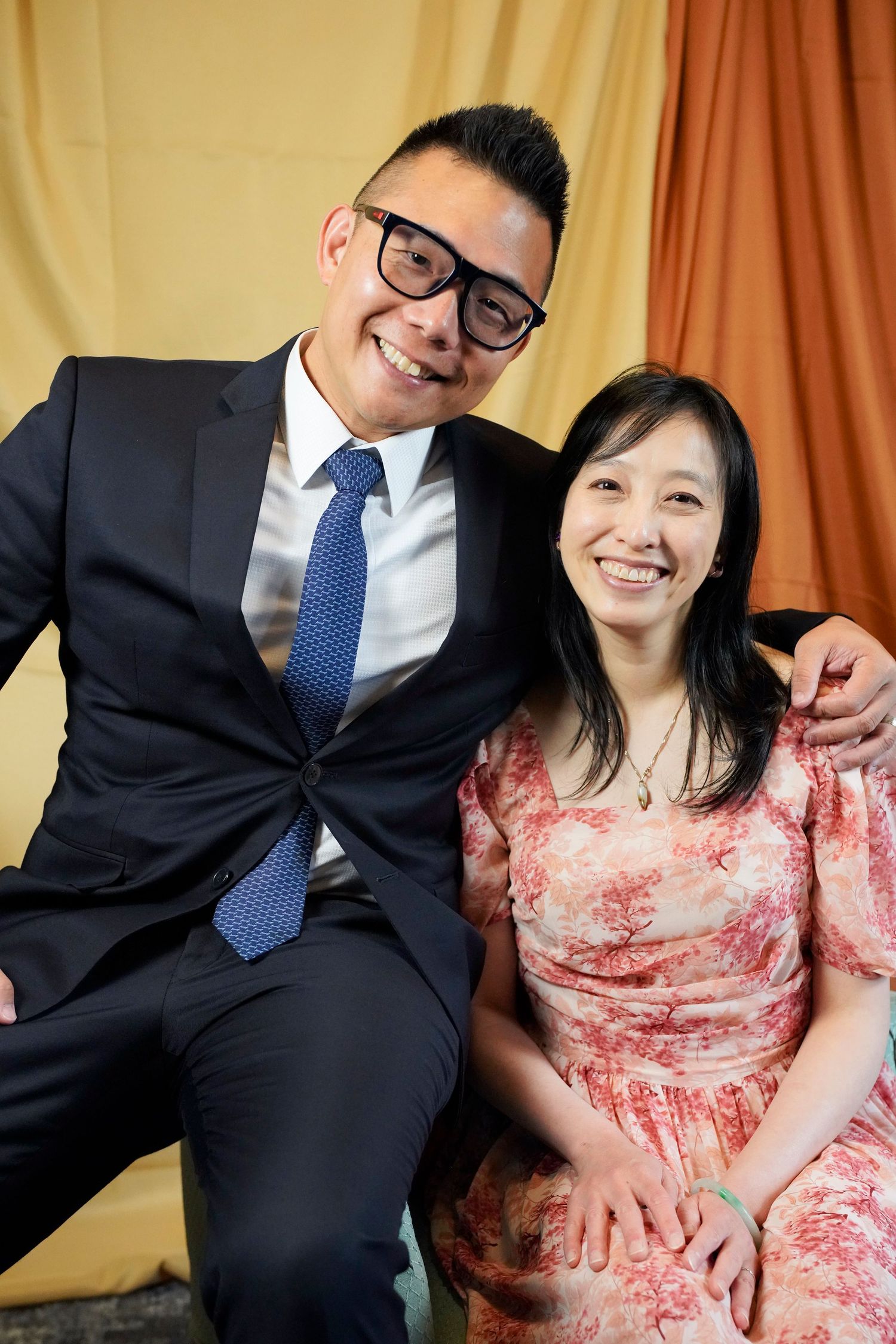 A couple sits together against a gold backdrop, with one wearing a suit and tie and the other in a floral pink dress.