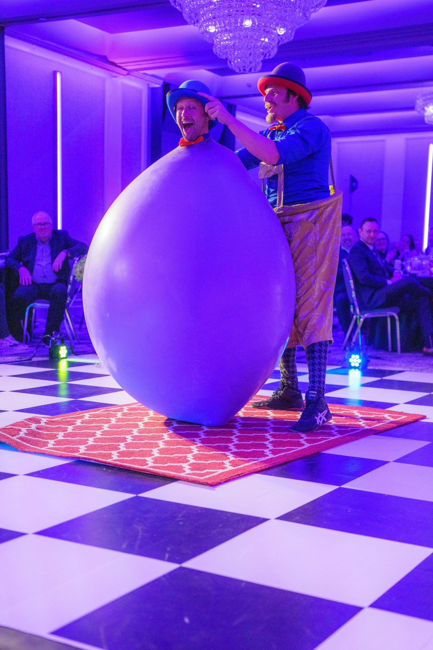 A performer in blue clothing stands next to a giant purple balloon on a checkered dance floor under purple lighting.