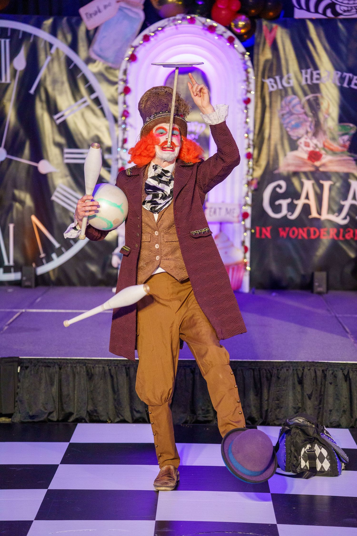 Performer in Victorian-style brown coat and vest juggles hats on stage with purple lighting and theatrical backdrop.