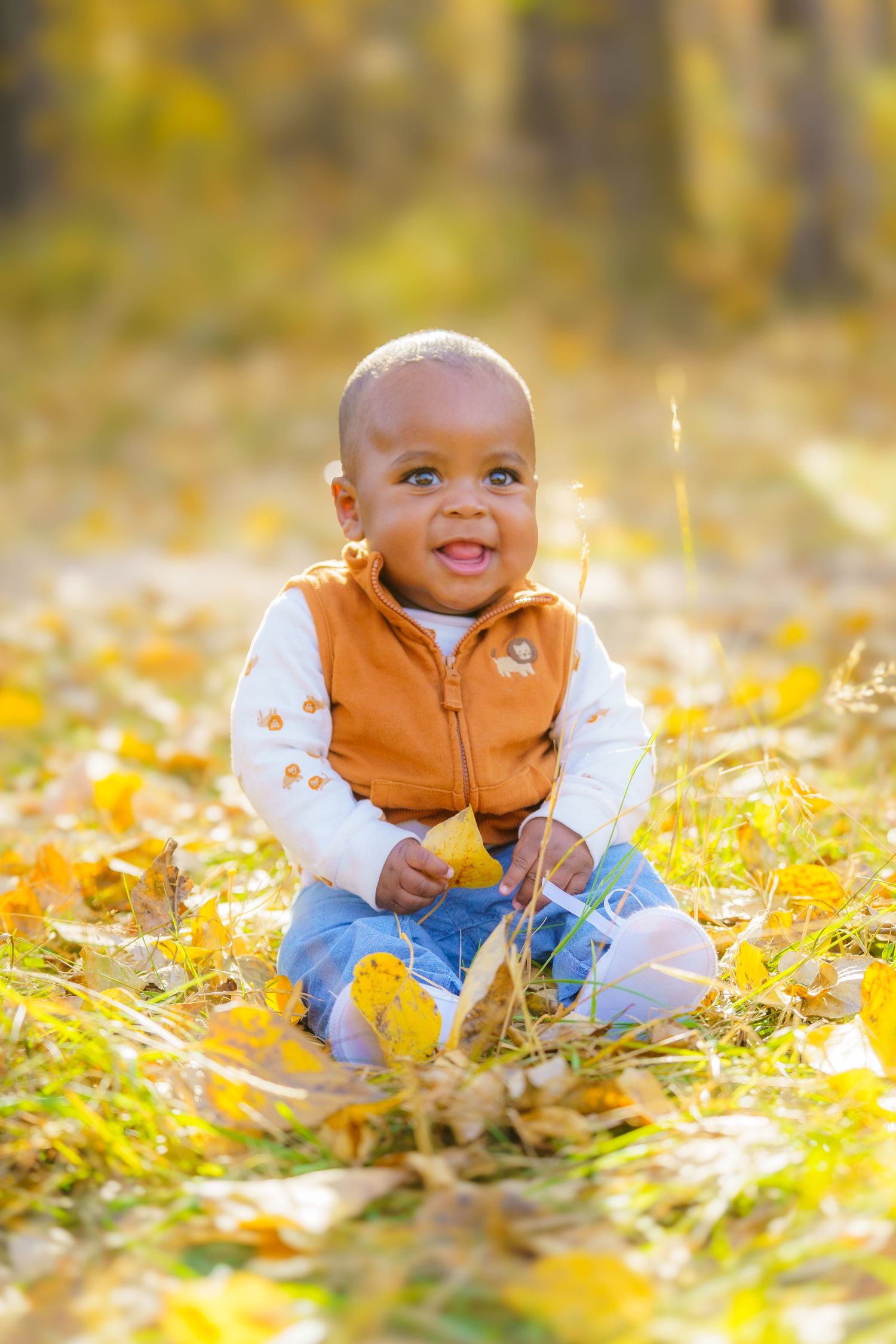 Small child wearing an orange vest sits happily among fallen yellow autumn leaves in golden sunlight.