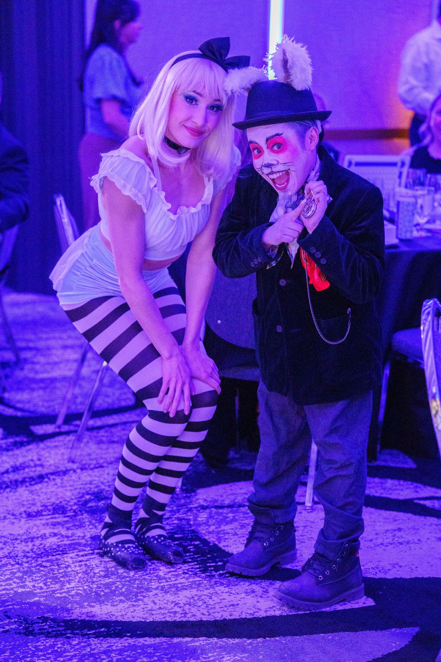 Two people in Halloween costumes pose together under purple lighting at what appears to be a costume party or event.