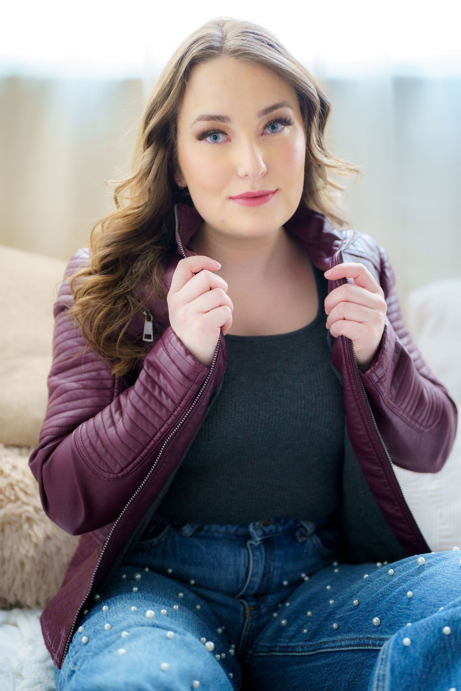 Woman in burgundy puffer jacket and jeans poses casually while sitting on couch with natural lighting.