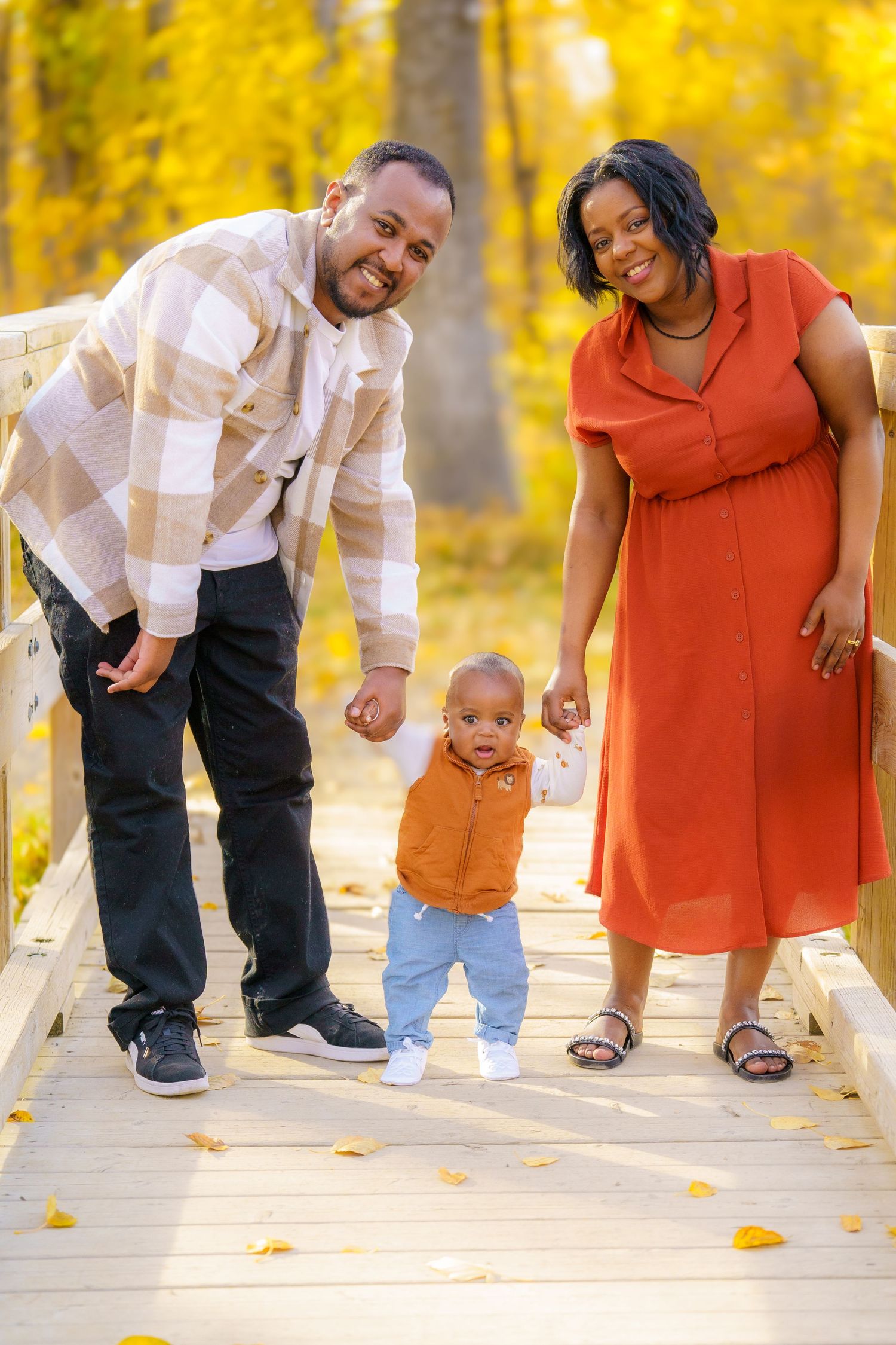Family in autumn attire walks together on wooden bridge surrounded by golden yellow fall foliage.
