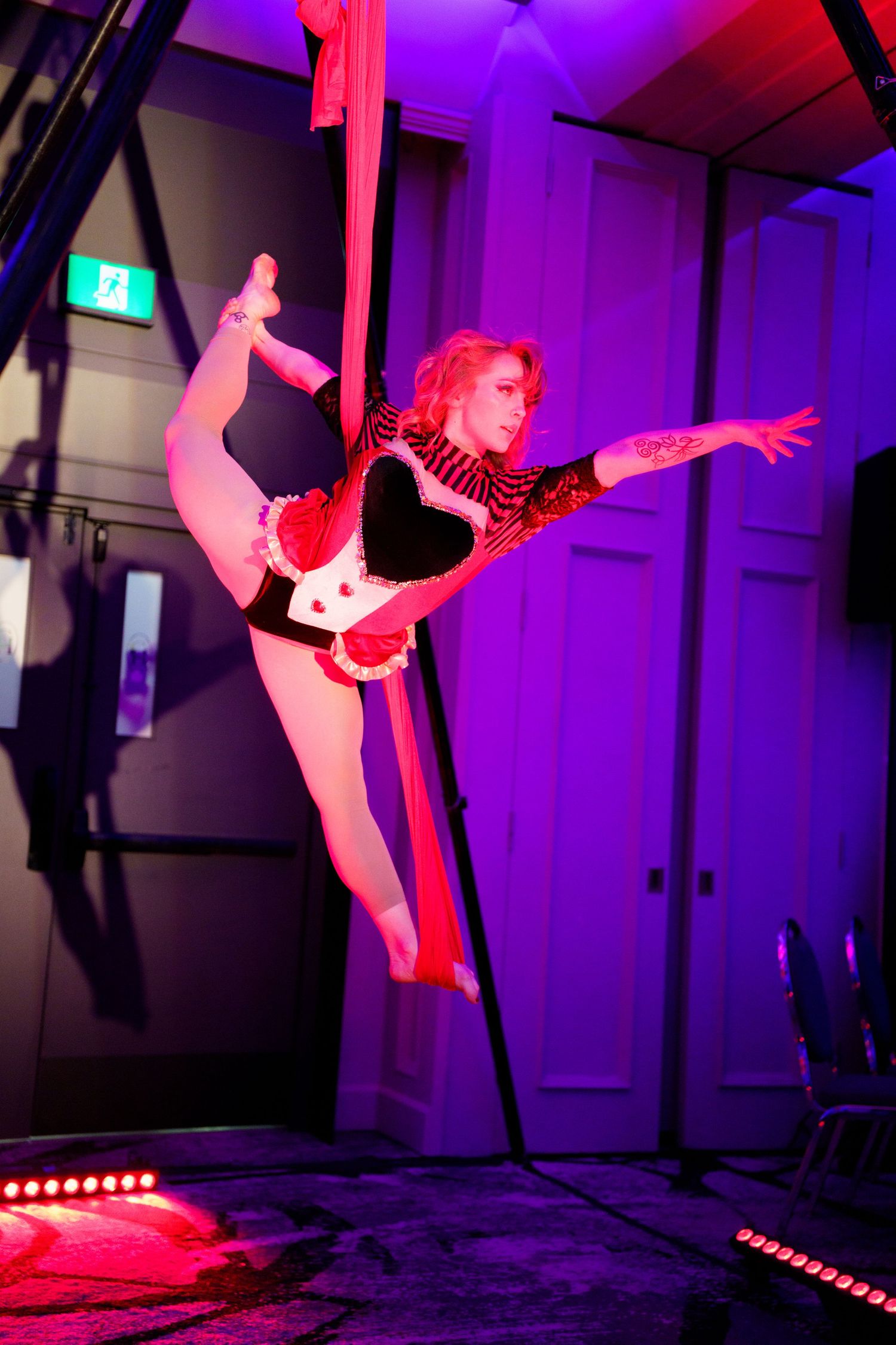 Acrobatic performer in red costume executes dramatic aerial pose on silks against purple-lit stage backdrop.