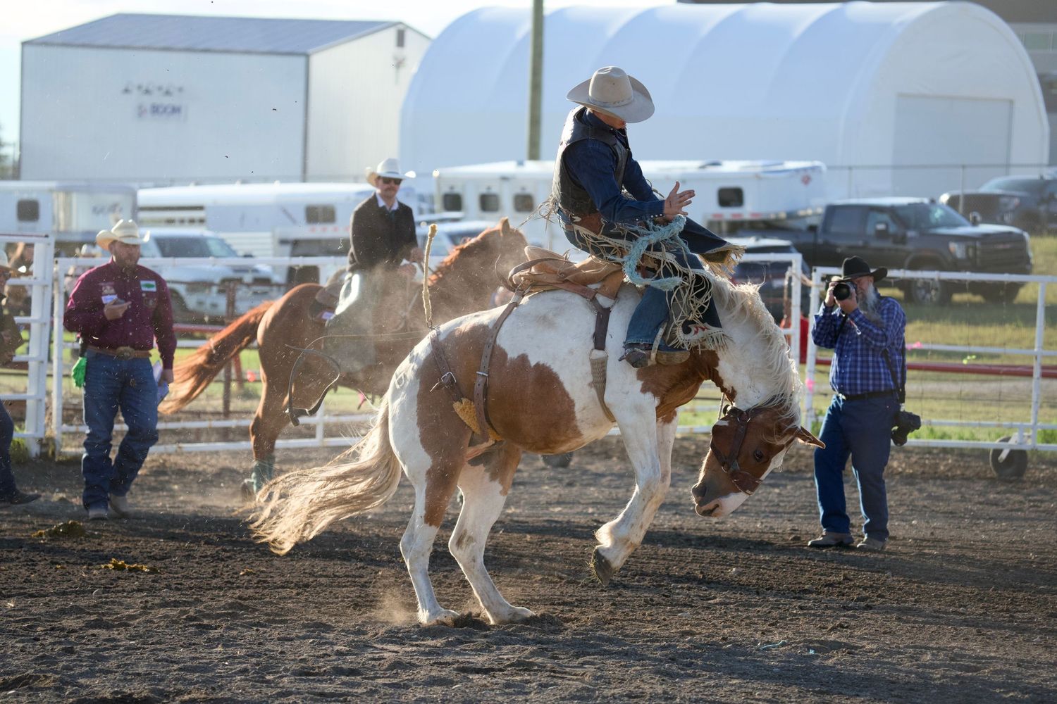 A rodeo rider competes on a bucking horse in an outdoor arena while handlers stand nearby.