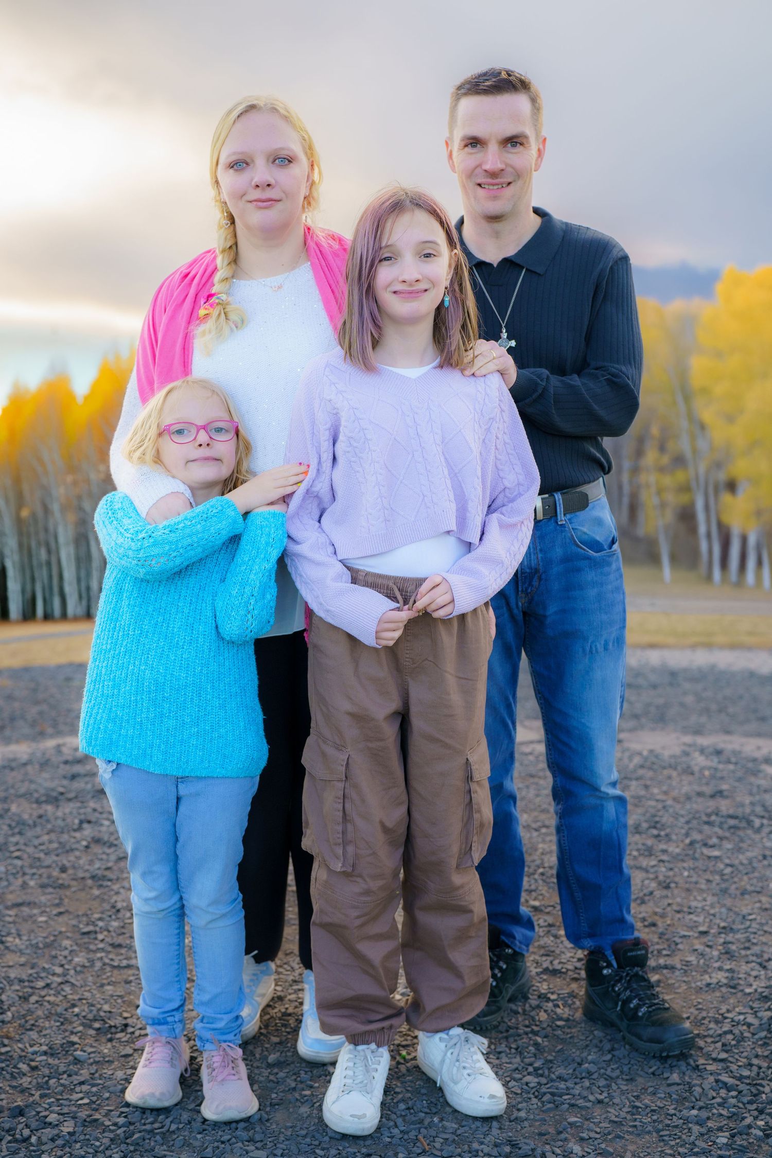 A family dressed casually poses together outdoors against an autumn forest backdrop with golden yellow trees.