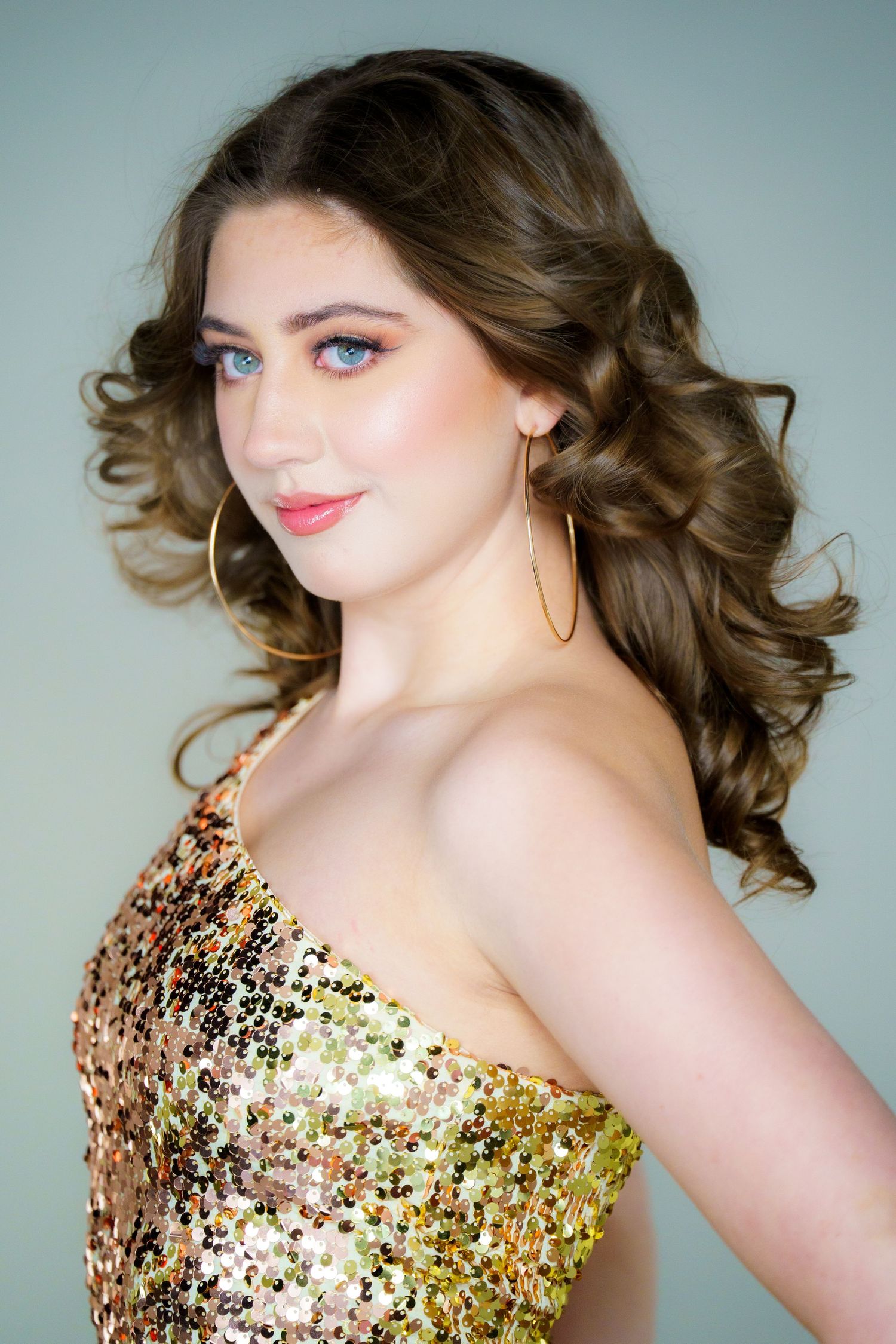 Professional portrait featuring a glamorous gold sequined dress, styled wavy brown hair, and elegant hoop earrings.