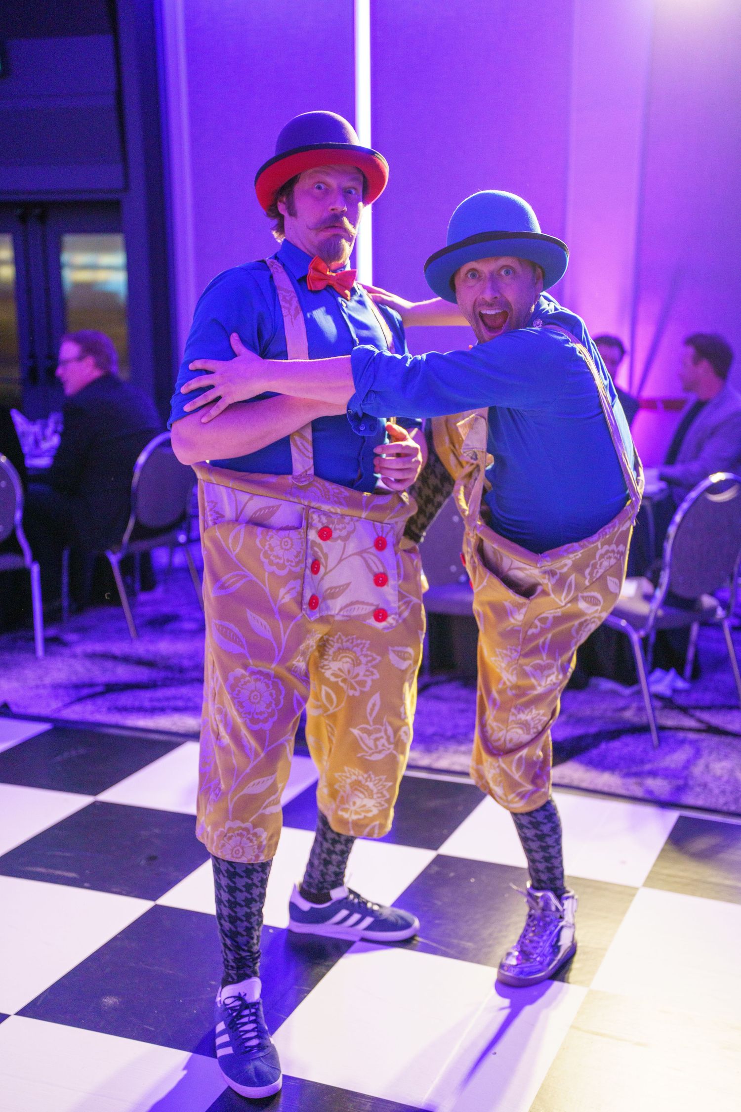 Two performers wearing bowler hats, colorful patterned pants and sneakers pose playfully on a checkered dance floor in purple lighting.