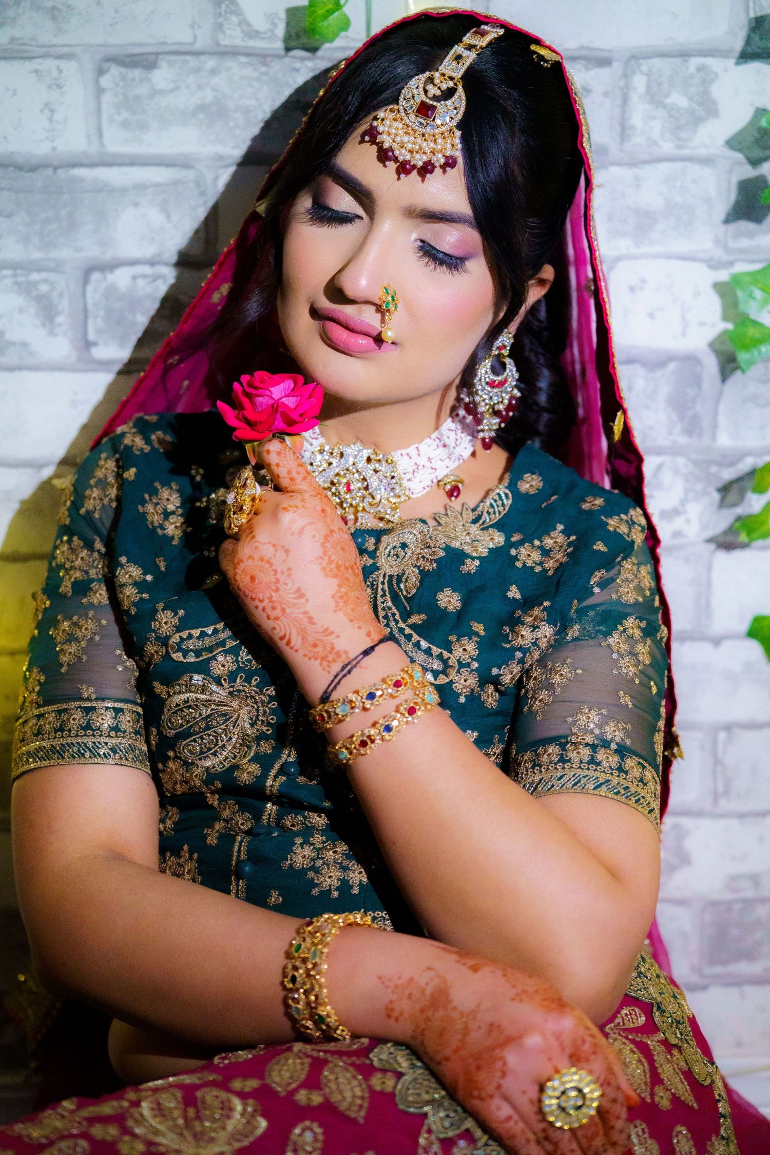 Traditional South Asian bride wearing teal and magenta wedding attire with gold jewelry and henna decorated hands holds pink rose.
