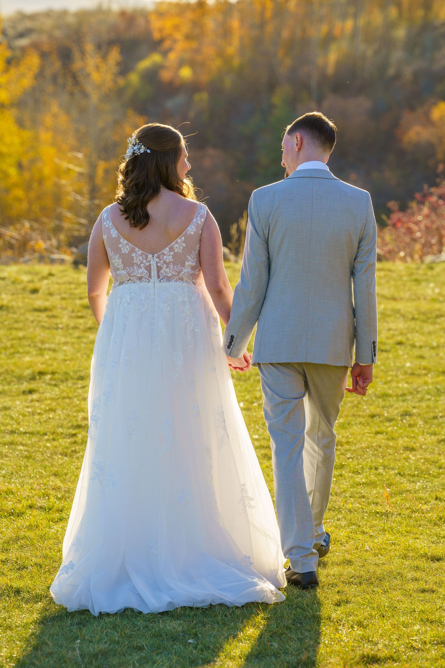 Newlywed couple walking on green grass during sunset with autumn foliage in background.