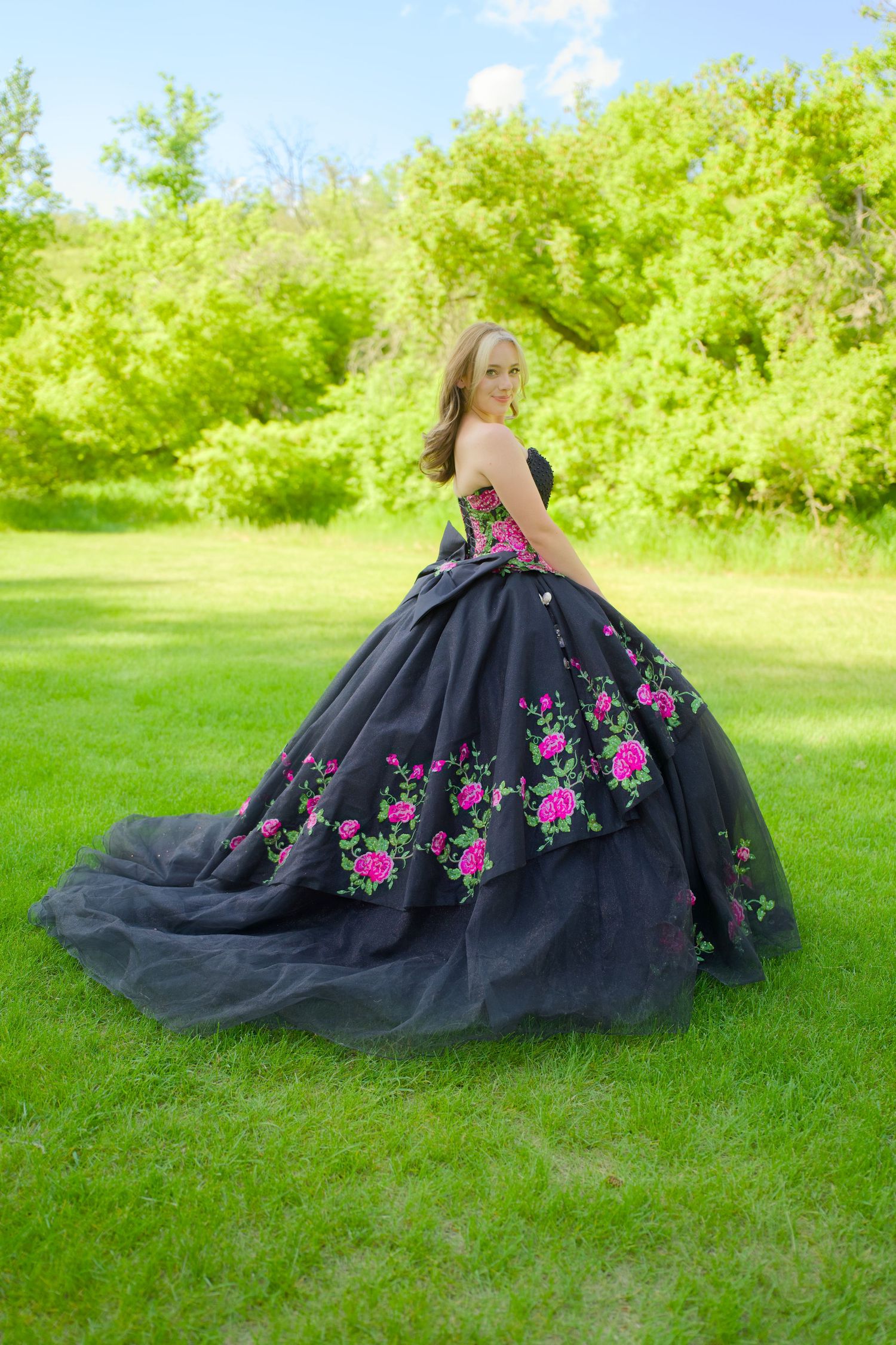 A black ball gown with pink floral embellishments is displayed on a lush green lawn with trees in the background.