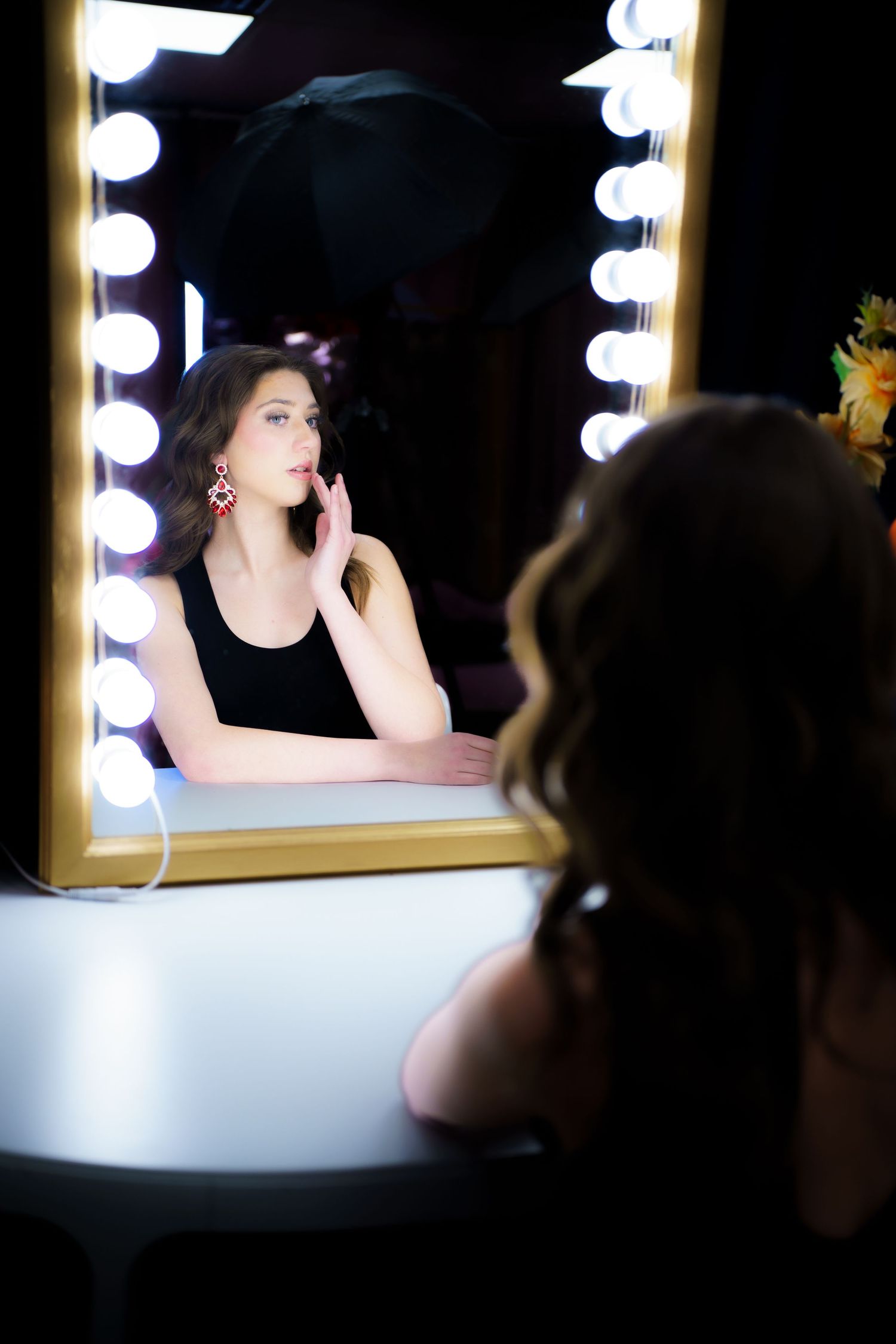 Someone looks in a well-lit makeup mirror with illuminated bulbs wearing a black tank top in a dark room with soft lighting.