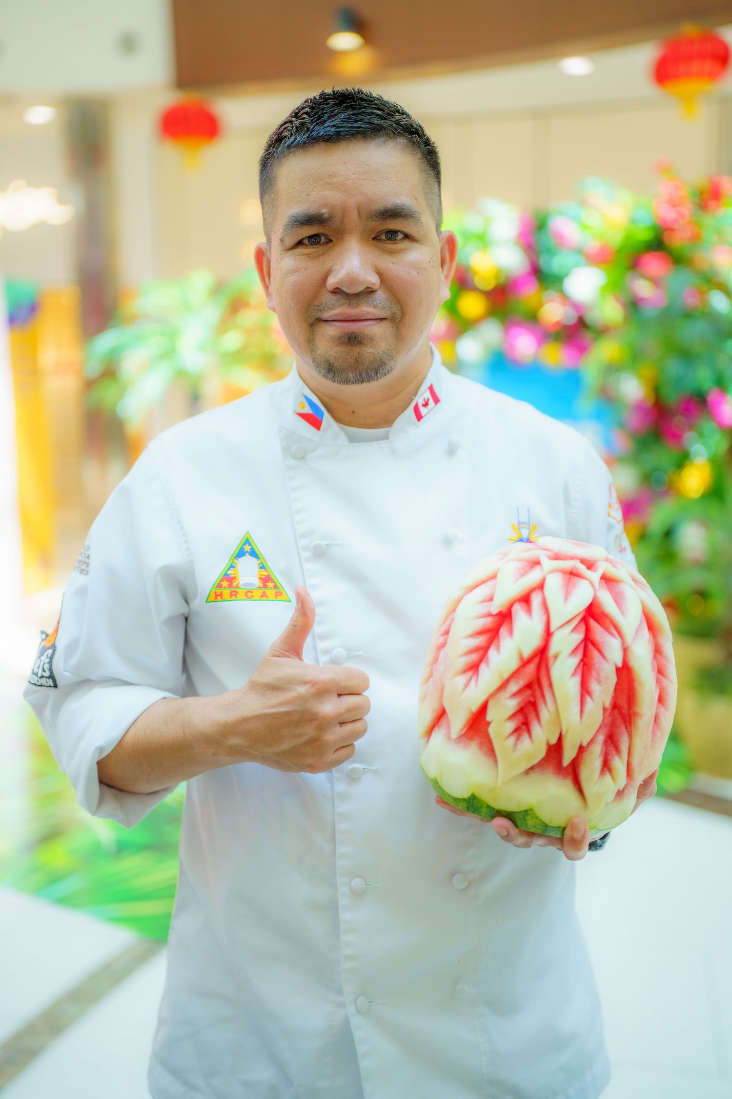 Chef in white uniform displays intricate watermelon carving with red leaf design in front of festive decor.