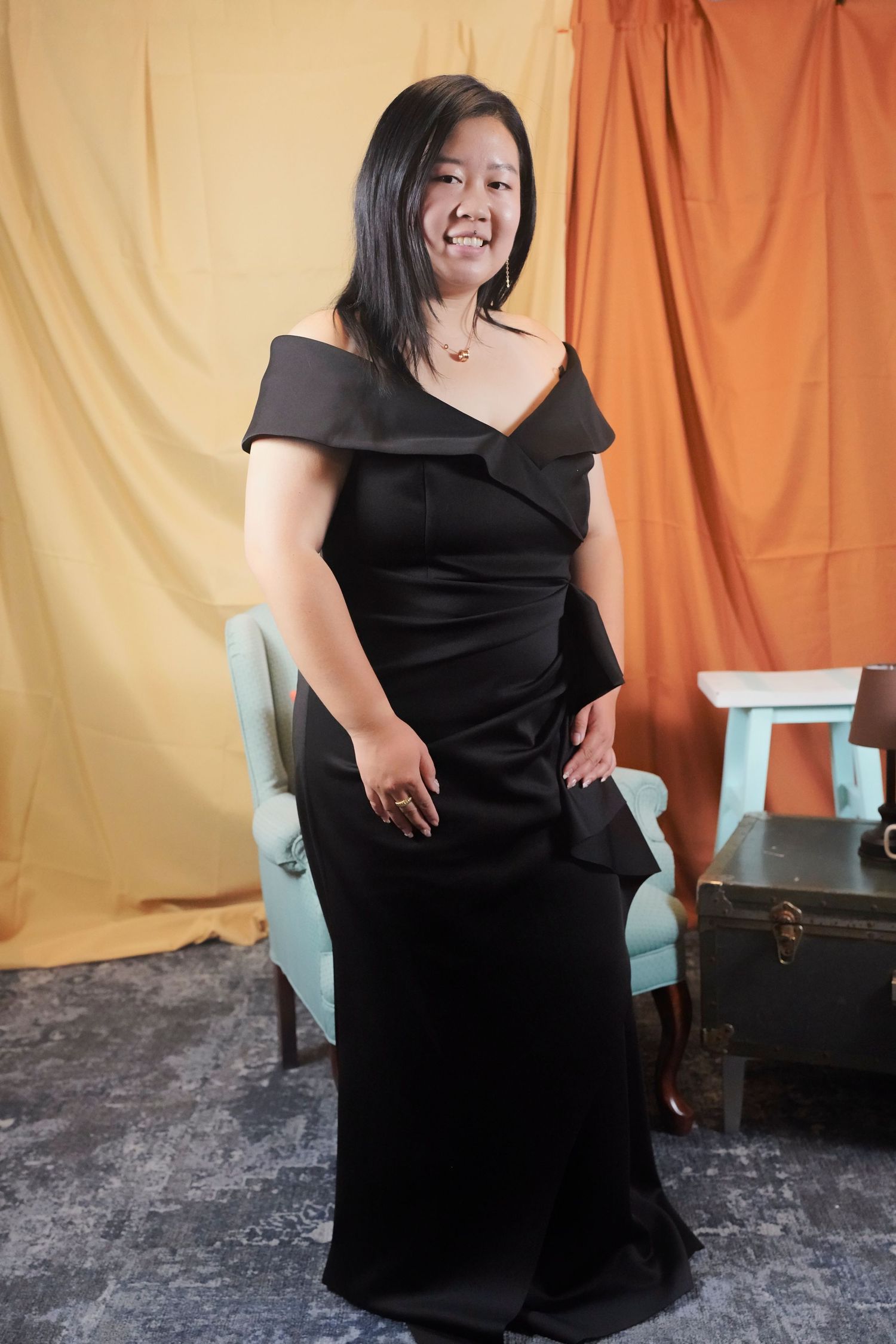 An elegant black off-shoulder floor-length evening gown is worn while posing in front of an orange and beige backdrop.