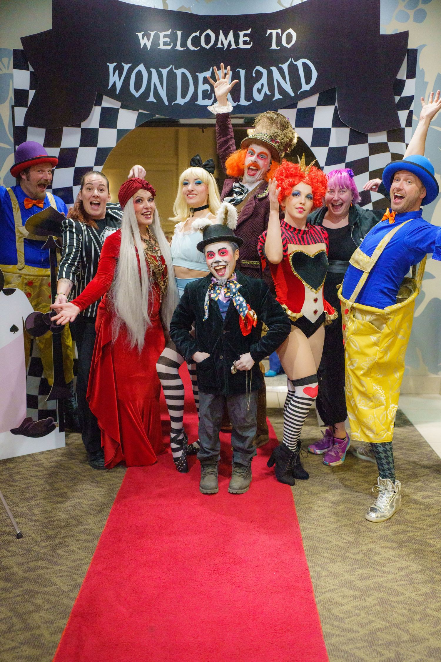 Costumed performers pose under Welcome to Wonderland sign with checkered border and red carpet beneath.