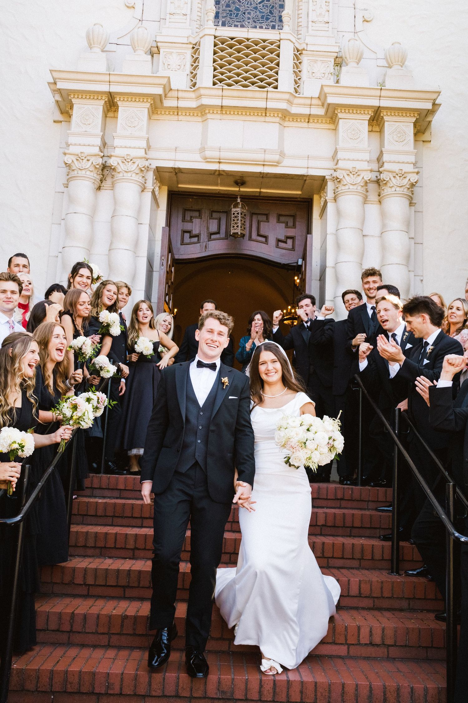Newlywed couple exits historic white church with bridal party in black attire gathering on brick steps.