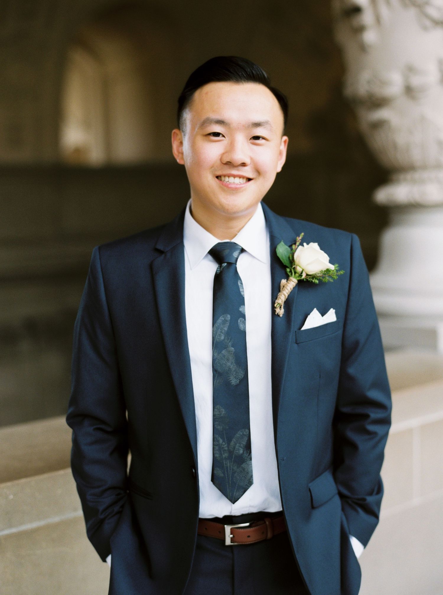 Groom in navy blue suit and tie with white boutonniere smiles while standing in formal archway.
