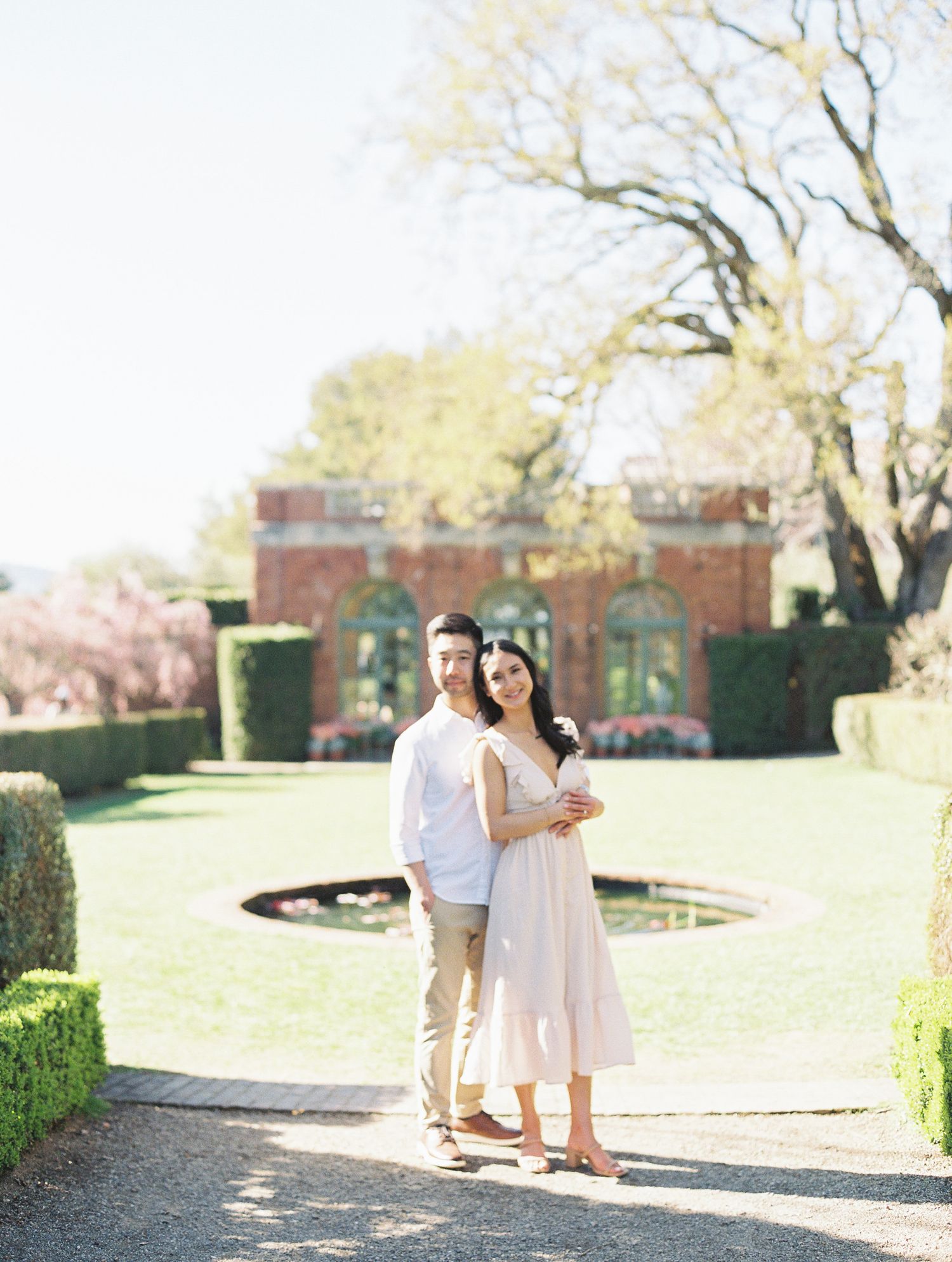 A couple in white outfits walks together through a sunlit garden path surrounded by blooming spring trees.