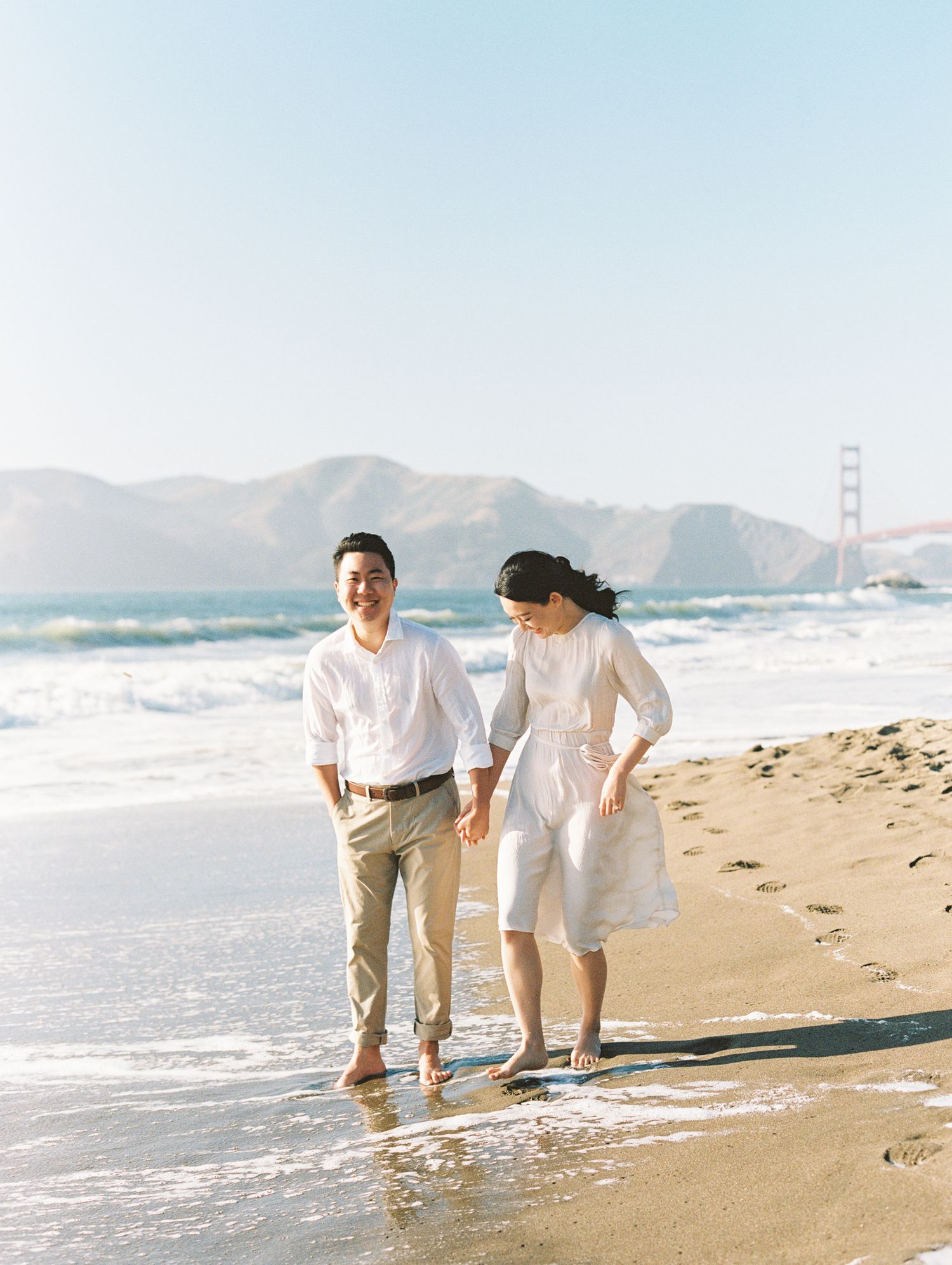A couple walks together on Baker Beach in San Francisco with the Golden Gate Bridge visible in the background.