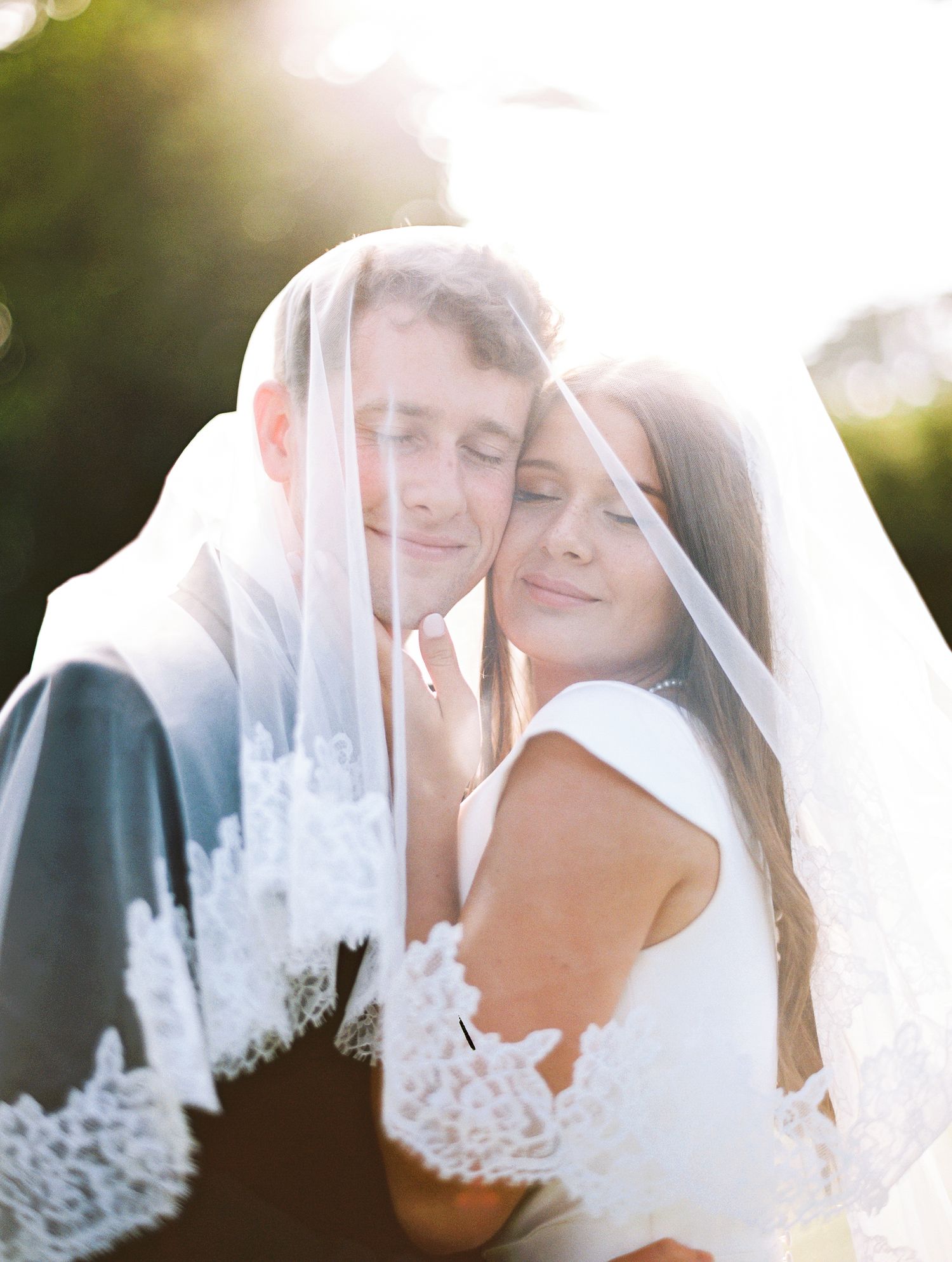 A romantic wedding portrait captured under soft sunlight with delicate lace veil details.