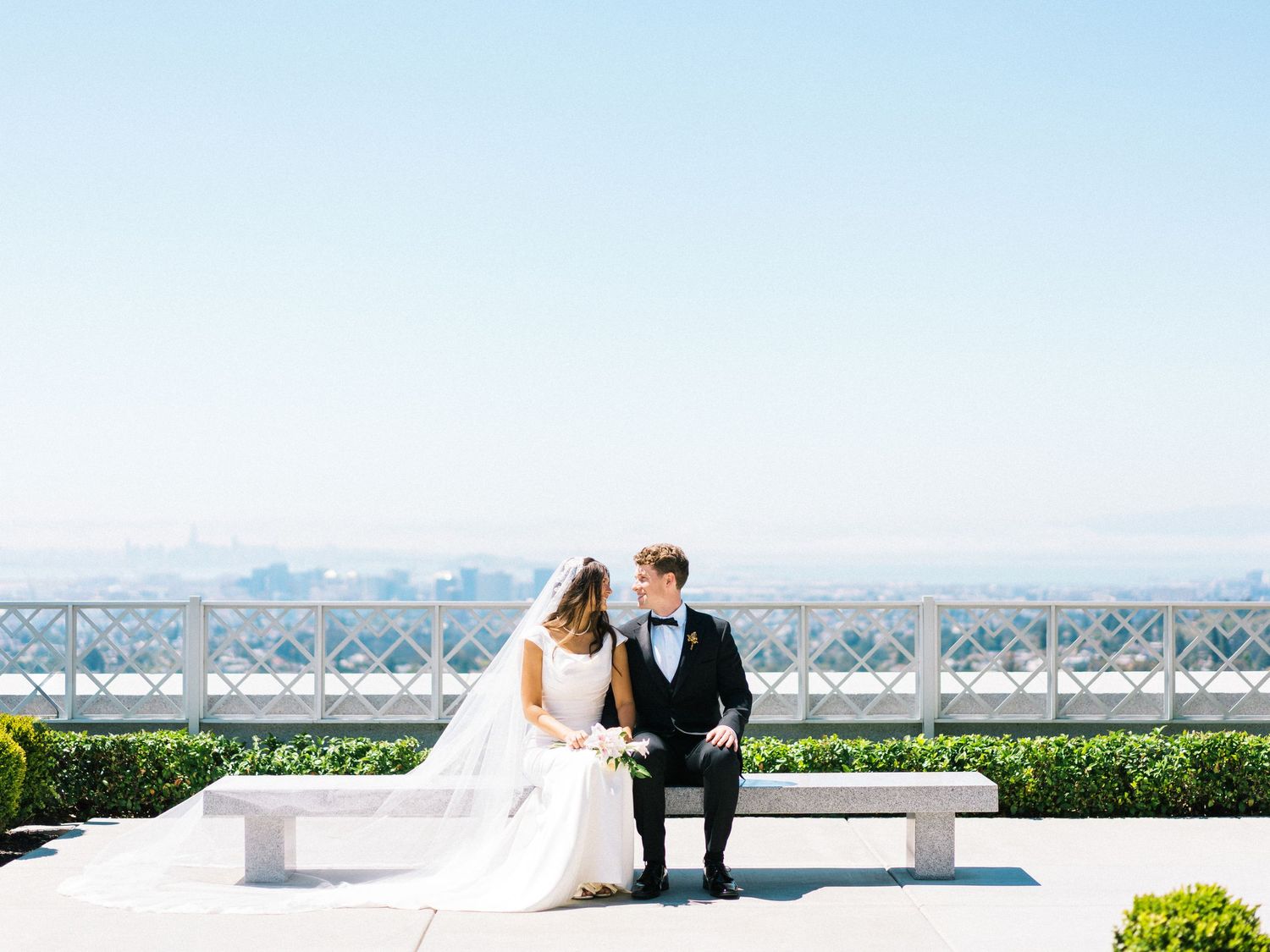 A couple in wedding attire sits together on a bench overlooking mountains and water on a sunny day.