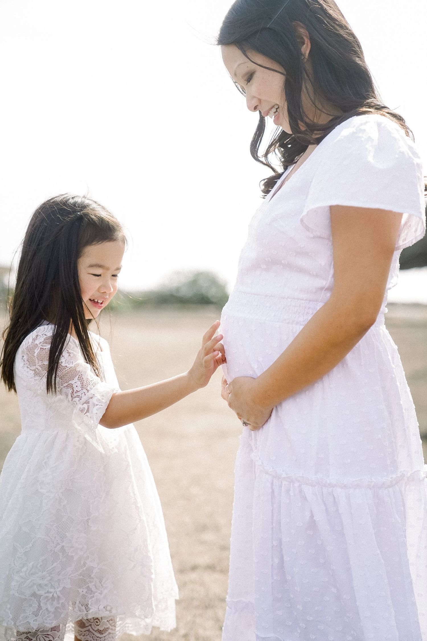 A young girl in a white lace dress touches her pregnant mother's belly while they share a tender outdoor moment.
