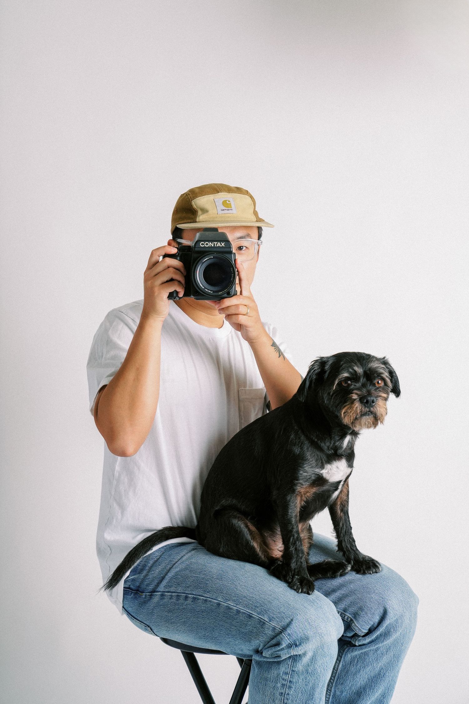 A person in a beige cap takes a photo while sitting with their black dog.