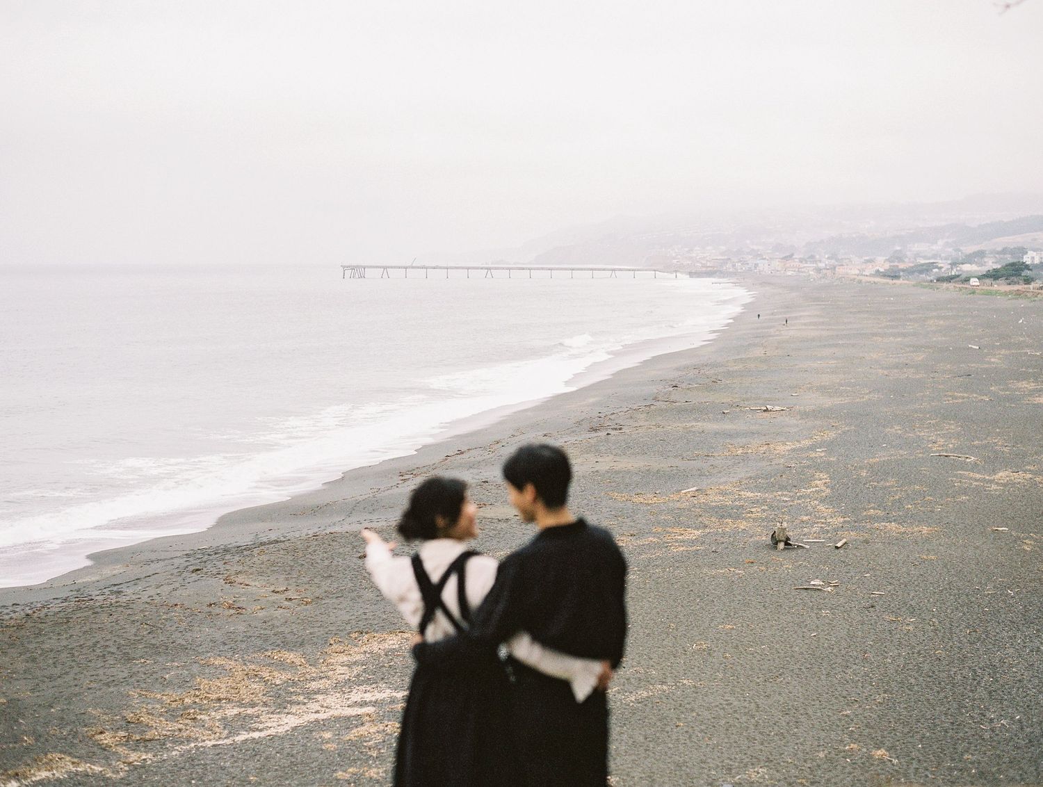 Two figures stand on a misty coastal beach overlooking the curving shoreline on an overcast day.