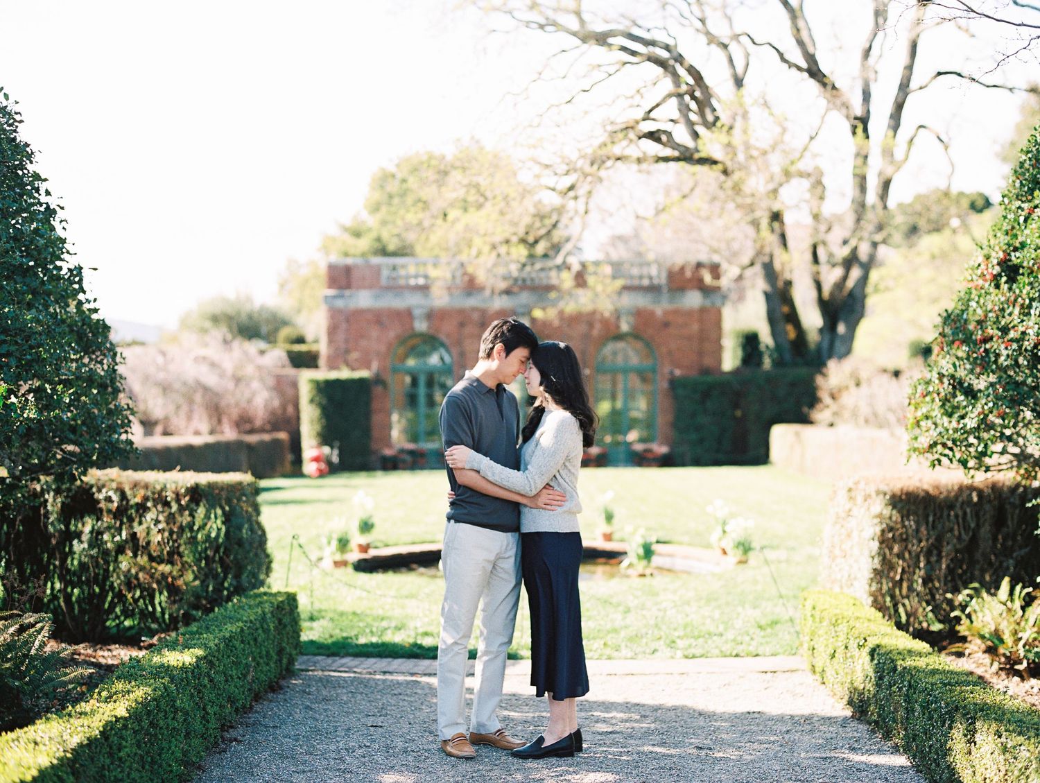 A couple shares a romantic moment on a garden path surrounded by manicured hedges and brick walls on a sunny day.