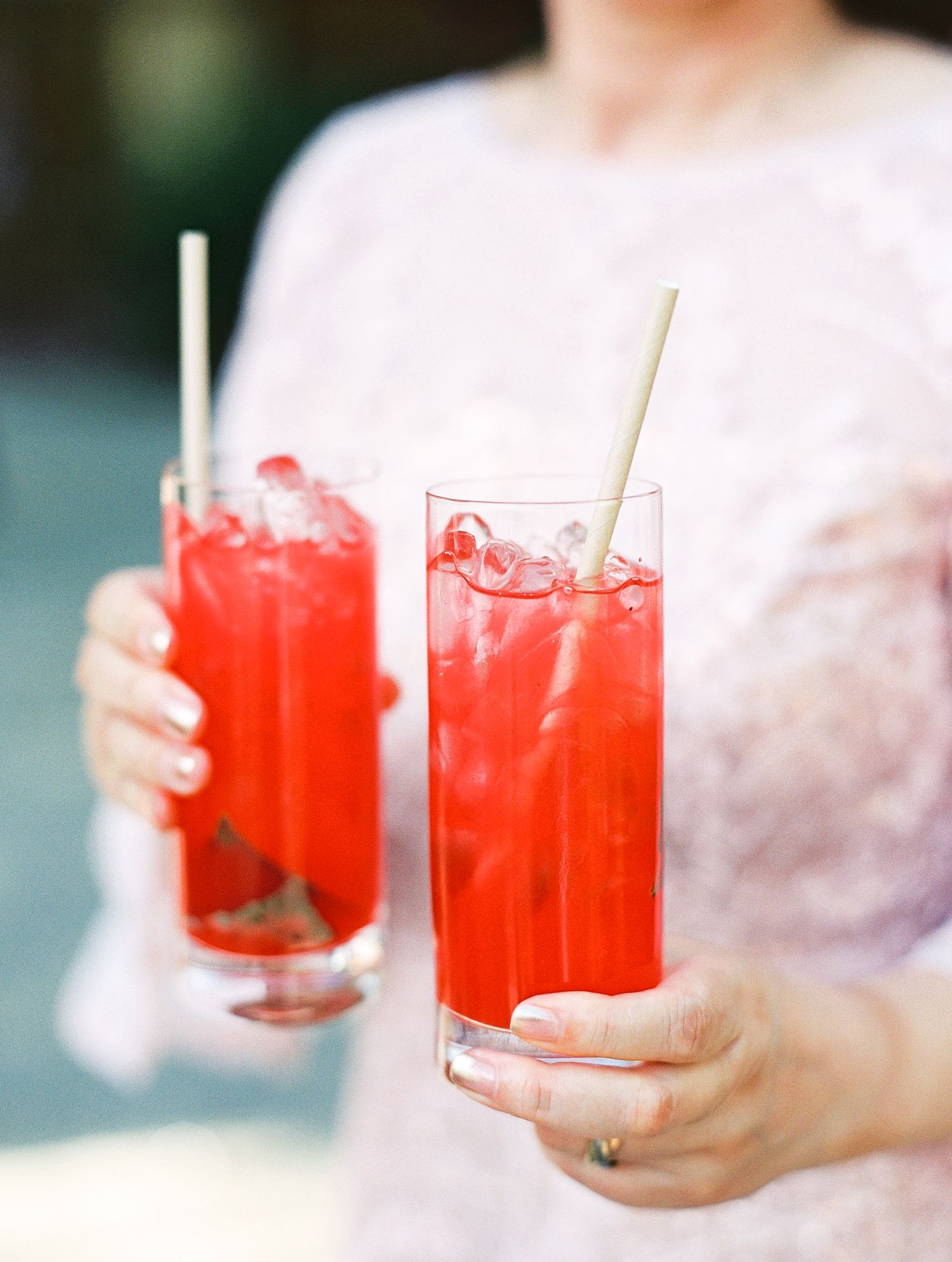 Two refreshing red cocktails with ice and straws are held in tall glasses against a light background.