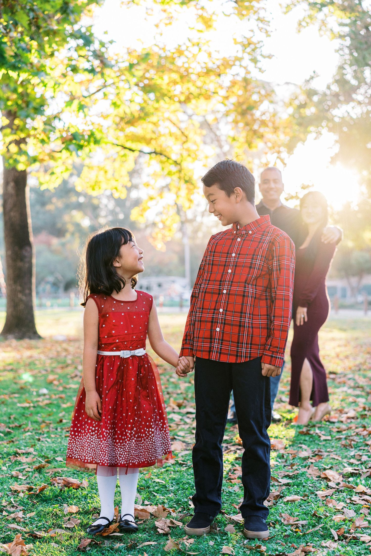 A young child in a red polka dot dress plays with family members among fallen autumn leaves in a sunlit park.