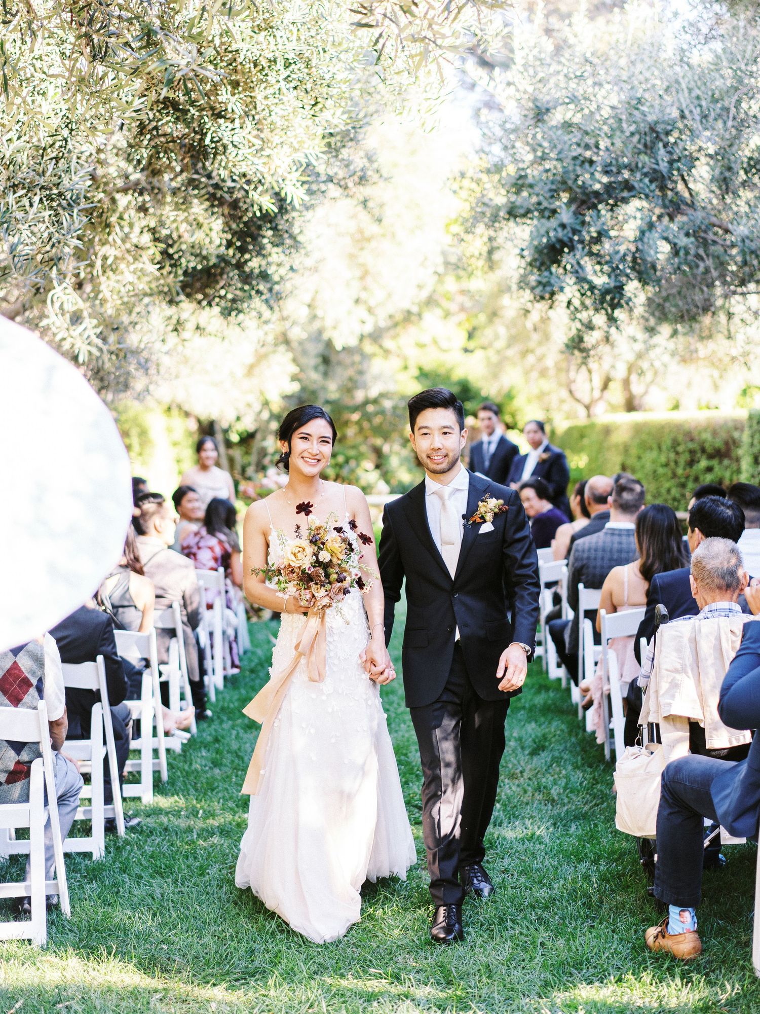 Happy couple walks back down the outdoor wedding aisle under trees while guests seated in white chairs look on.
