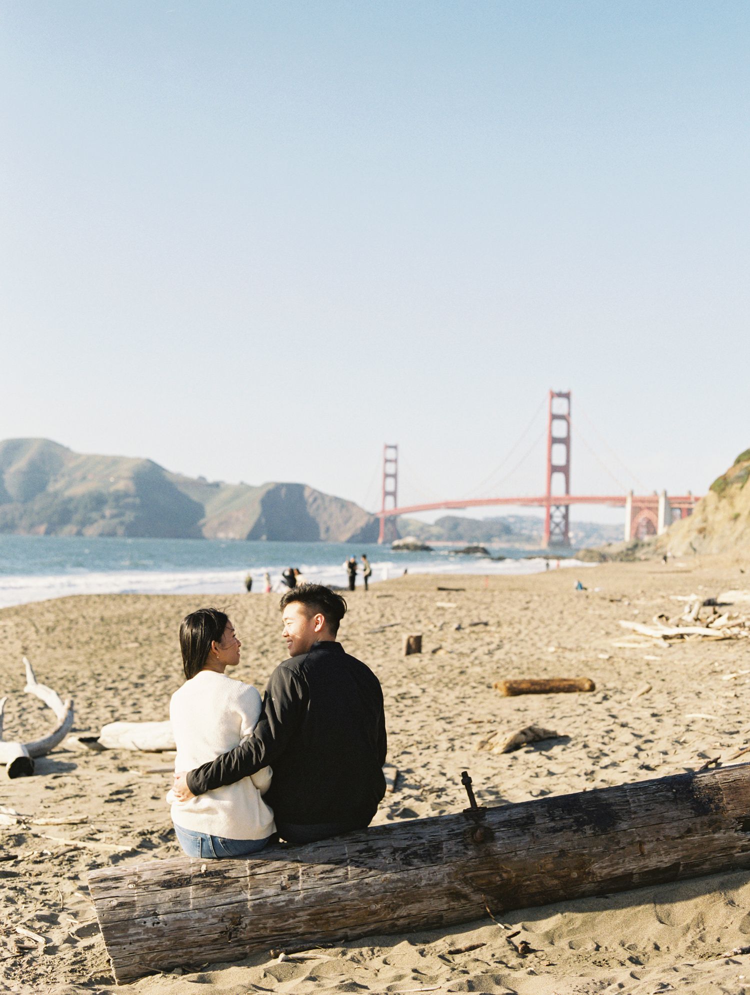 A couple sits on a driftwood log at Baker Beach with the iconic Golden Gate Bridge in the background on a sunny day.